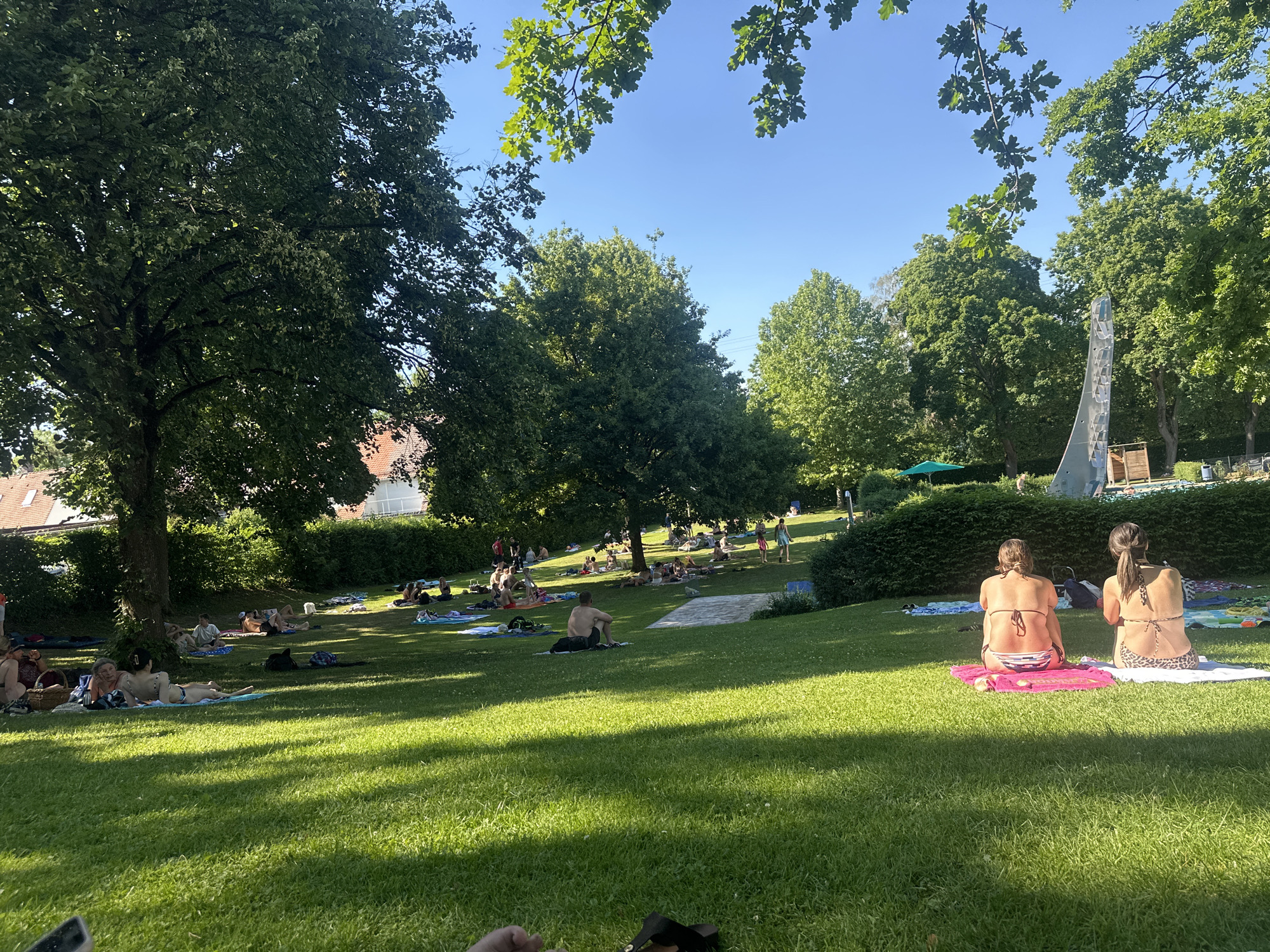 The image shows a sunny day at the swimming pool (Freibad) in Mainburg, Germany, with many people relaxing on the grassy area. In the foreground, two women are sitting on towels, facing away from the camera, wearing bikinis. The grass is green and well-maintained, with dappled sunlight and shadows from the surrounding trees. In the middle ground, numerous individuals are spread out on towels and blankets, sunbathing, sitting, or lying down. Some are in swimwear, while others are clothed. A few trees provide shade, and a white paved area is visible in the distance. The background features more trees, some buildings with red-tiled roofs, and a blue sky. A sculptural element or fountain is partially visible on the right side of the image, and a green umbrella is set up further back. The overall scene depicts a relaxed summer atmosphere at an outdoor public swimming facility.