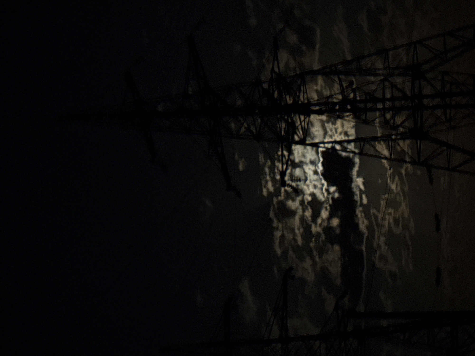 This is a dark, nocturnal image featuring the silhouette of a large, intricate metal lattice structure, likely an electricity pylon or a bridge truss, against a moonlit, cloudy sky. The structure, composed of numerous dark, crisscrossing beams and supports, spans much of the right and central portions of the frame, creating a strong foreground element. Its geometric patterns are visible despite the overall darkness.
Behind the dark structure, the sky is illuminated by the moon, which shines through and reflects off dense, textured clouds. These clouds appear in various shades of grey and bright white, forming a mottled, almost abstract pattern that contrasts sharply with the deep black of the foreground elements. The left side of the image is predominantly dark, hinting at an expansive night sky.
There is no discernible activity or event taking place; the scene is static. The time of day is clearly night, and the weather appears to be cloudy. The presence of the large industrial structure suggests a setting outside of a natural landscape, possibly near infrastructure. The overall mood conveyed is one of stark contrast, mystery, and an almost dramatic quietude. No text is visible anywhere in the image.