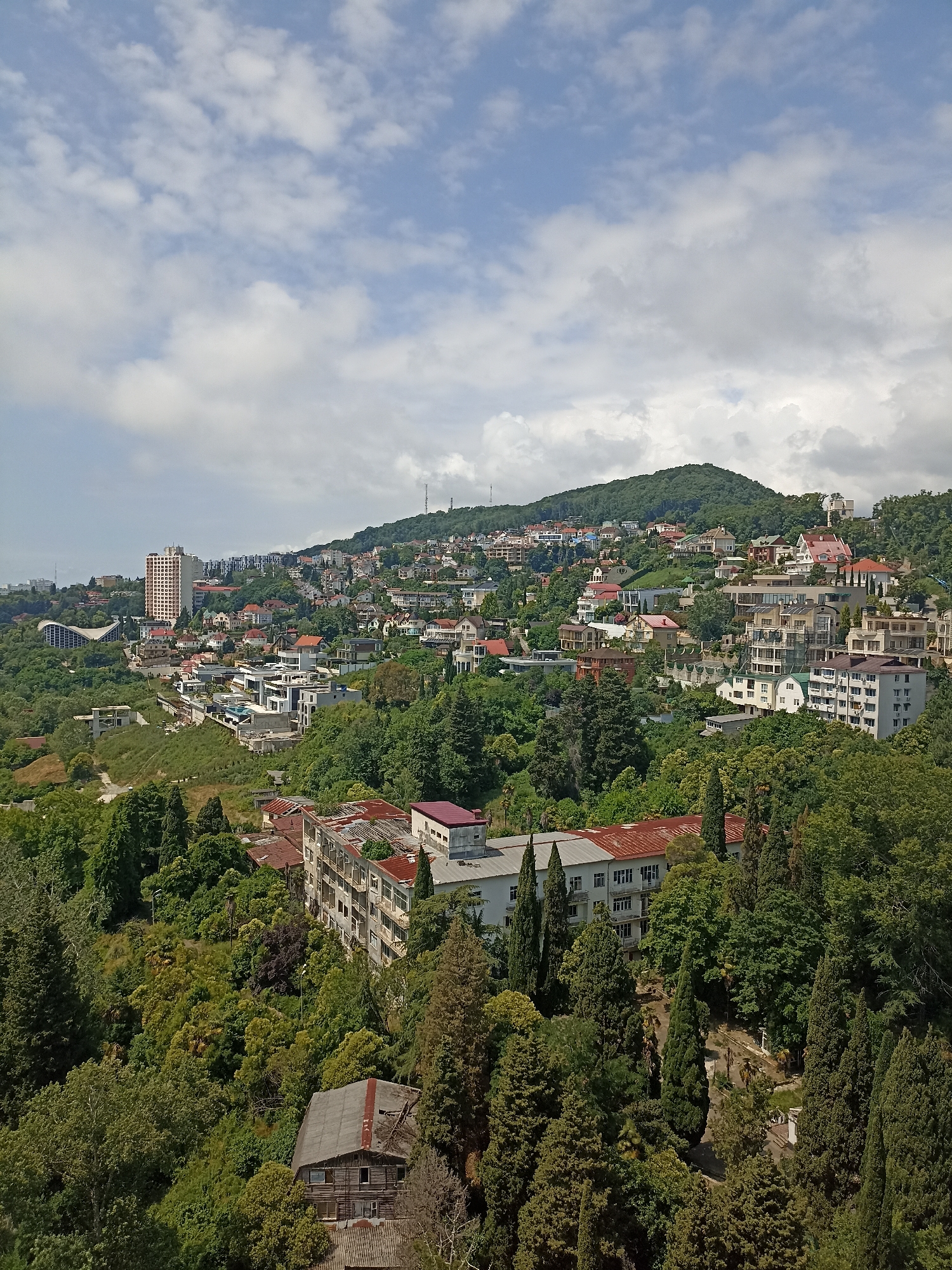 The image depicts a dense hillside residential area with a mix of older and modern buildings, overgrown with lush green trees. In the background, a tall, light-colored apartment building stands out, and further up the hill, a network of streets and houses climbs towards a verdant mountain peak. A telecommunications tower is visible on the far right of the mountain. The foreground features more trees and some dilapidated structures, suggesting that parts of the area might be in disrepair or undergoing development. The sky is partly cloudy with white cumulus clouds scattered across a pale blue expanse. The scene appears to be a panoramic view of a coastal city nestled against a mountainous backdrop, likely Sochi, Russia, given its characteristic topography and architecture. There are no visible people or specific actions taking place.