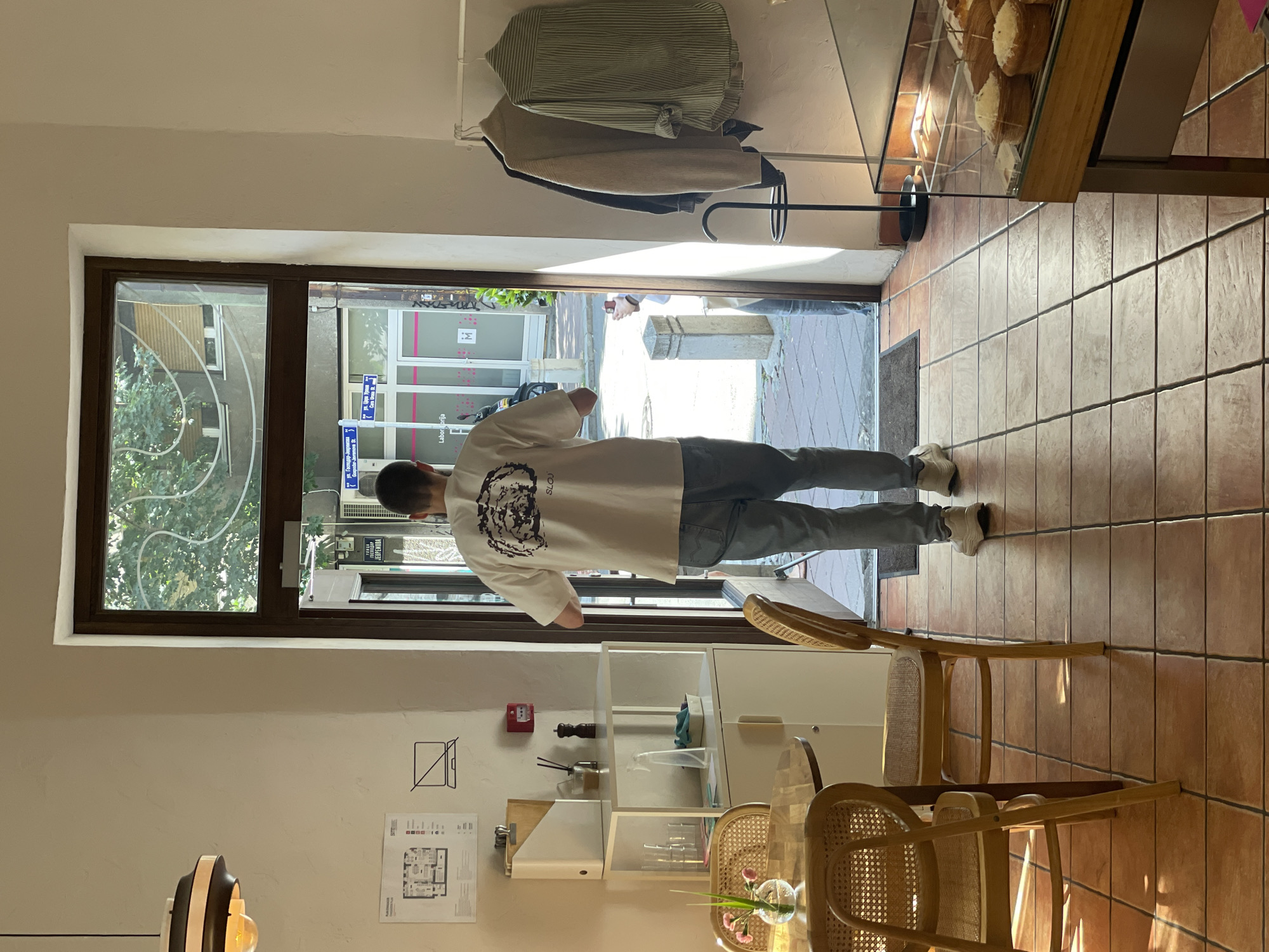 A man with short dark hair walks away from the camera towards an open doorway in a cafe. He is wearing a light-colored t-shirt with a dark graphic on the back and light blue jeans. The cafe has terracotta tile floors and wooden chairs with woven seats and backs. A white shelving unit with a few items on it is to the left of the doorway. Outside the doorway, street signs are visible, including one with Cyrillic text that reads "ул. Георги Димитров" (Georgi Dimitrov Street) and another that reads "ул. Цара Душана" (Tsar Dusan Street), indicating the location is in Belgrade, Serbia, likely at the intersection of these two streets. The cafe has a display case with baked goods on the right side of the image, and clothing items hang on a rack near the doorway. Sunlight streams in from the doorway, casting shadows on the floor.
