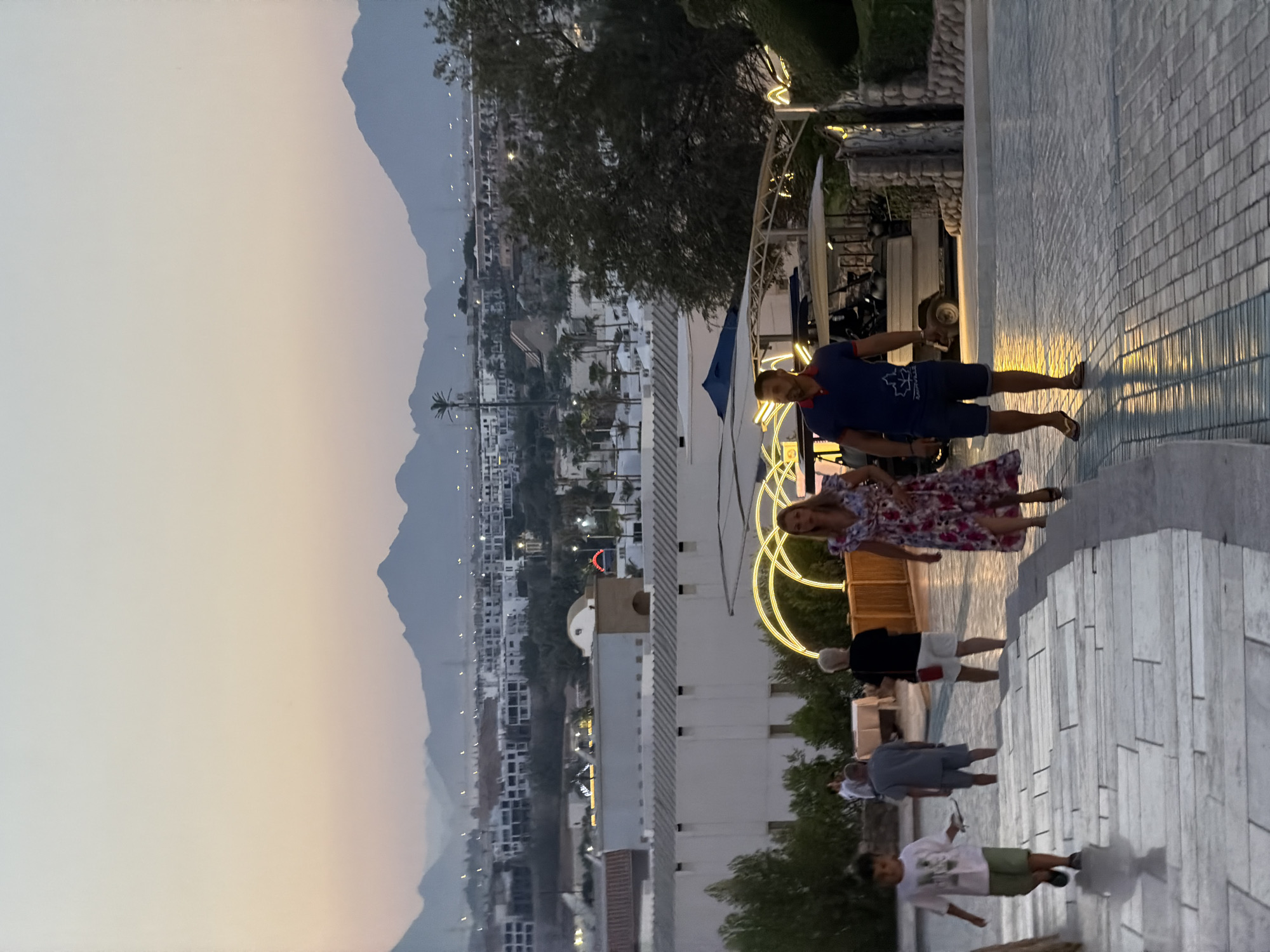 In Sharm El Sheikh, Egypt, an outdoor scene unfolds at dusk with a soft, pastel sky transitioning from pink to pale blue. A tiered landscape features a wide stone staircase in the foreground, leading down towards a paved walkway. Several people are visible navigating this descent. On the left, a young boy in a white t-shirt and green shorts walks down the stairs. Further down, a man in a gray t-shirt and shorts walks upwards, and a woman in a black top and white shorts also moves upwards. In the mid-ground, a couple is descending the stairs; the woman wears a floral print dress, and the man is in a blue polo shirt and dark shorts. Behind them, a golf cart is parked near a structure with a blue awning. The middle ground showcases buildings with white facades, interspersed with palm trees and illuminated by decorative yellow arches. In the distance, a city sprawls across the landscape, backed by a range of imposing mountains under the fading light. The overall atmosphere suggests a resort or residential area in Sharm El Sheikh, likely towards the evening.