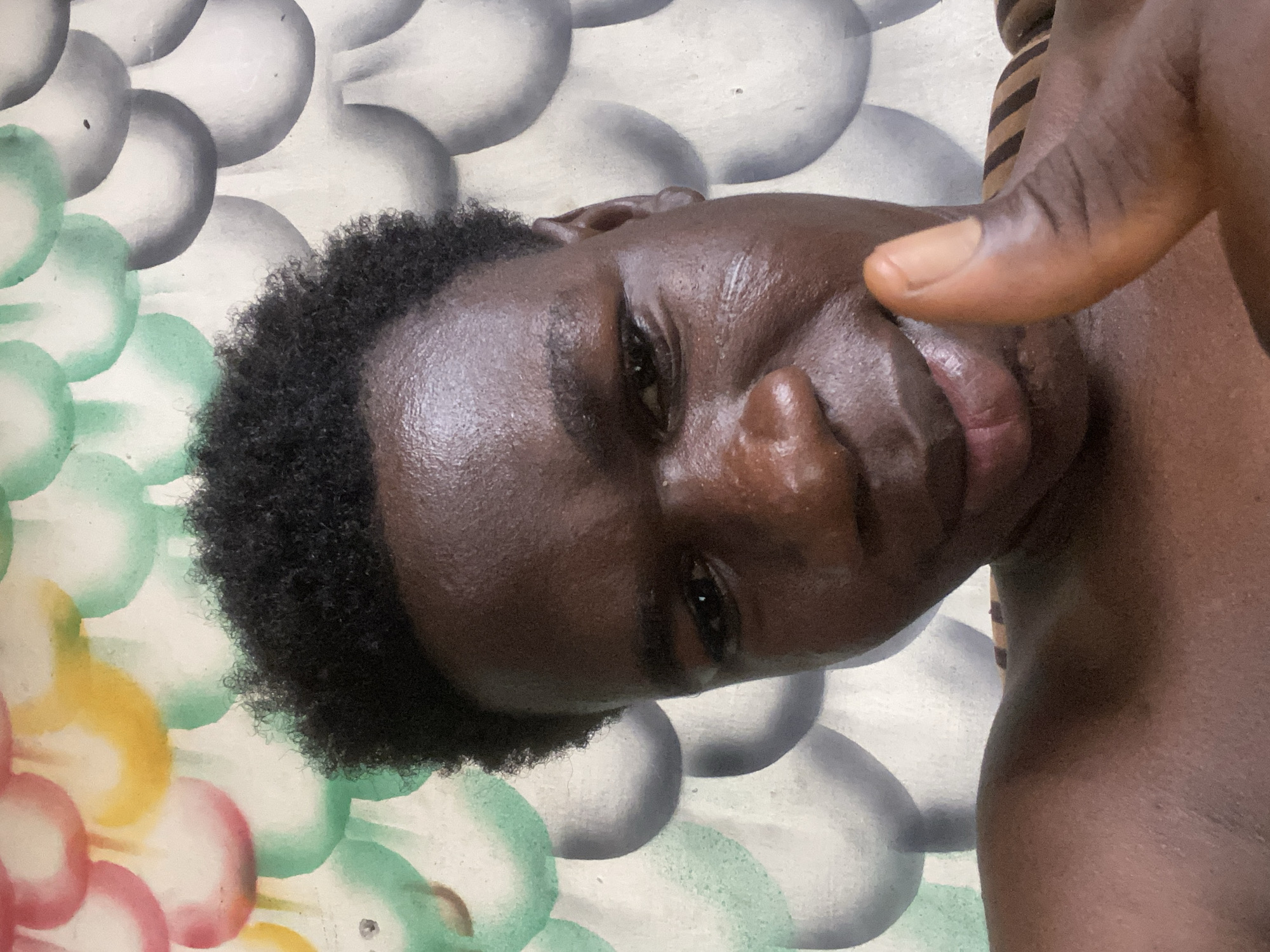 A close-up, slightly angled shot of a dark-skinned person with short, dark, curly hair. The person's face is glistening, possibly due to sweat or moisture. Their eyes are partially closed, and their mouth is slightly open. A dark-skinned hand with a lighter-toned thumb is positioned near the person's mouth. The background features a textured surface with rounded shapes in shades of grey, white, green, yellow, and pink. No specific information within the image confirms its location within Mararaba, Nigeria.
