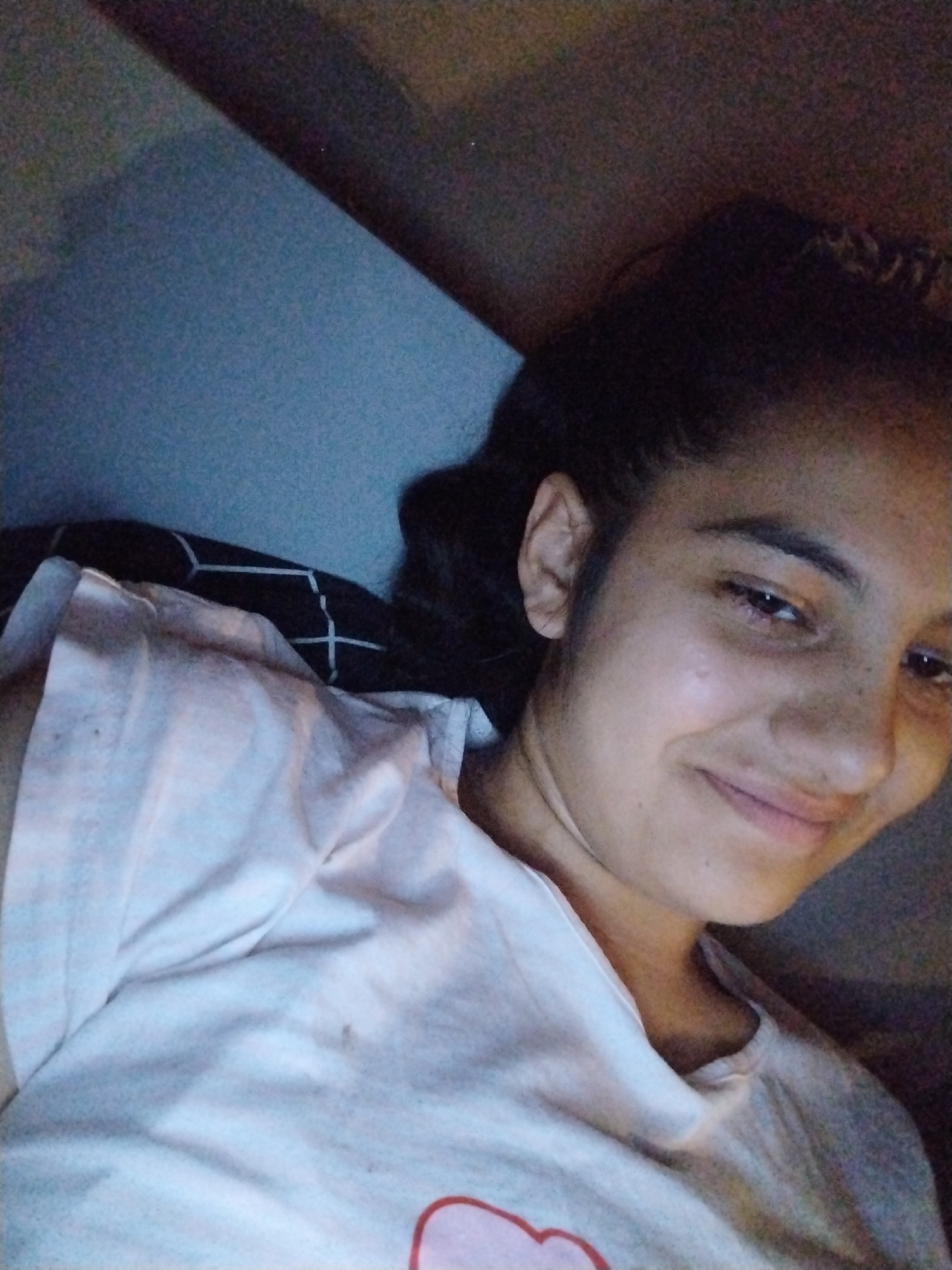 A close-up, overhead shot shows a young woman with dark hair pulled back, smiling gently as she looks into the camera. She is wearing a light-colored t-shirt. She is lying down, possibly on a bed, with a black and white patterned pillow visible behind her. The background is dark and blurry, suggesting an indoor setting, likely at night. The lighting is dim, casting soft shadows on her face.