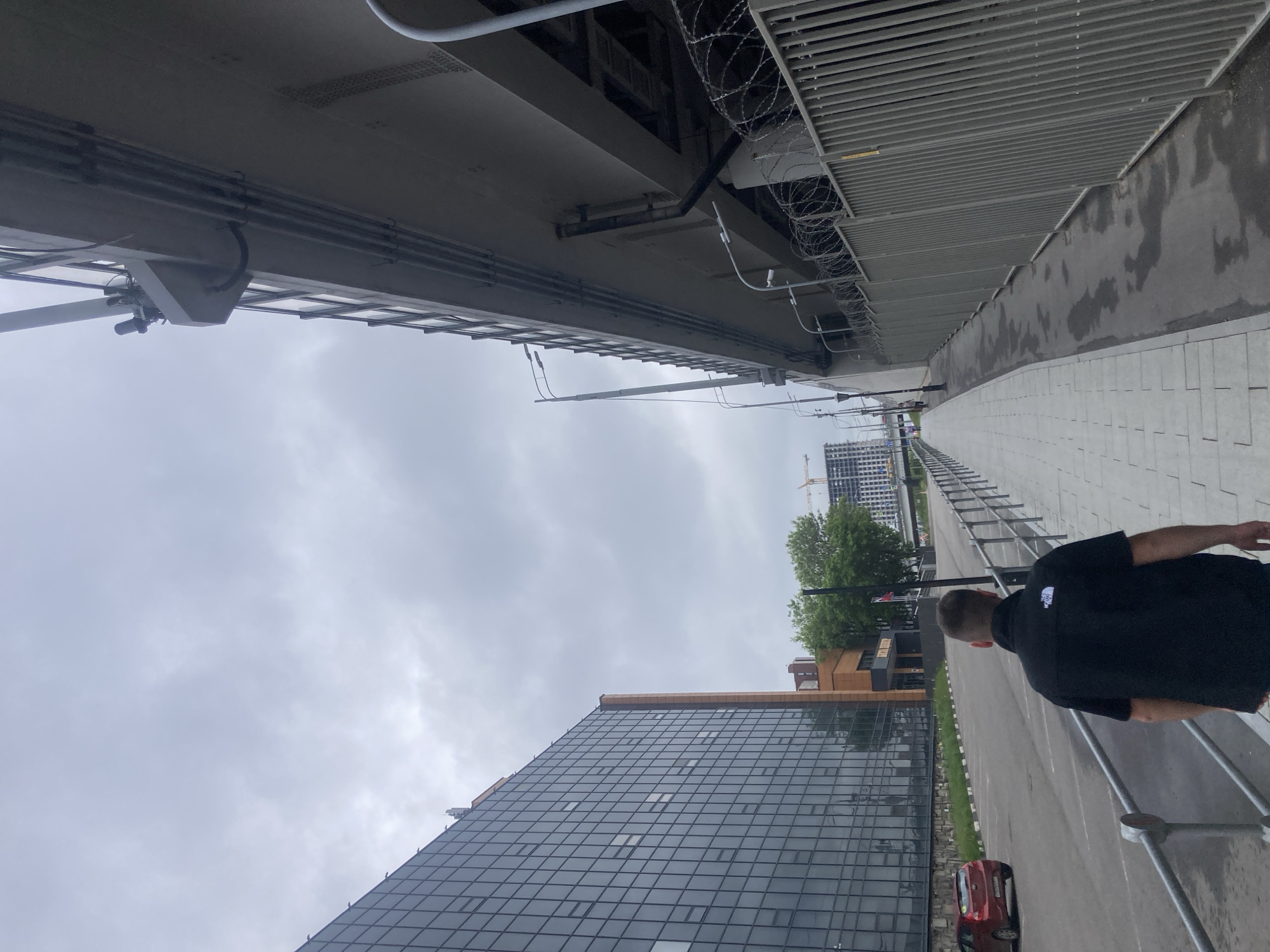 The image presents a view from below a large overpass or bridge structure, capturing the surrounding urban environment in Khimki, Russia. The sky is overcast with a heavy cloud cover.
On the left side of the frame, the underside of the bridge is visible, showcasing a complex network of pipes, cables, and support structures. A security system, possibly including cameras and barbed wire, is in place along the edge of the bridge.
To the right of the bridge, a sidewalk is lined with gray pavers. A road runs parallel to the sidewalk, with a pedestrian wearing a black t-shirt on the right side of the frame.
Across the road is a modern glass-facade building with a reflective exterior. Next to it, a shorter building has a facade made of wood. Trees are situated behind the buildings.
In the distance, construction cranes and high-rise buildings hint at ongoing urban development in the area. A red vehicle is parked along the road.