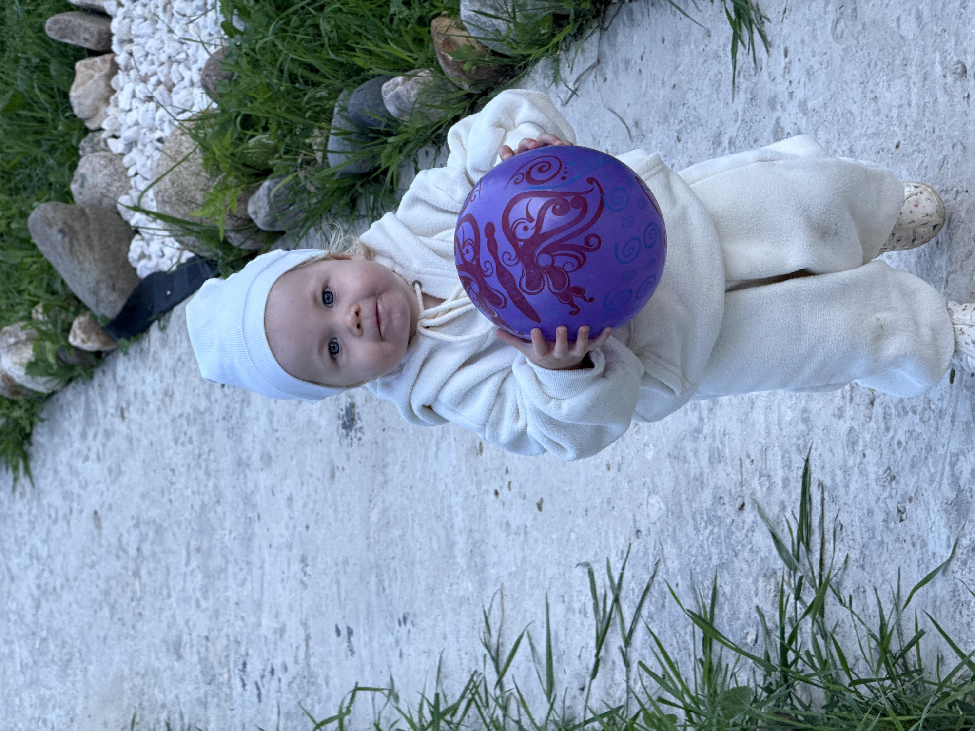 A toddler wearing a white hat and cream-colored fleece clothing stands on a concrete path in an outdoor setting, likely a garden or park. The toddler is holding a purple ball with a decorative pattern. To the right of the path, there is a border of rocks and grass. The scene is set in Ryzhkovo, Russia, as indicated by the surrounding environment which suggests a rural or suburban location consistent with the provided context.