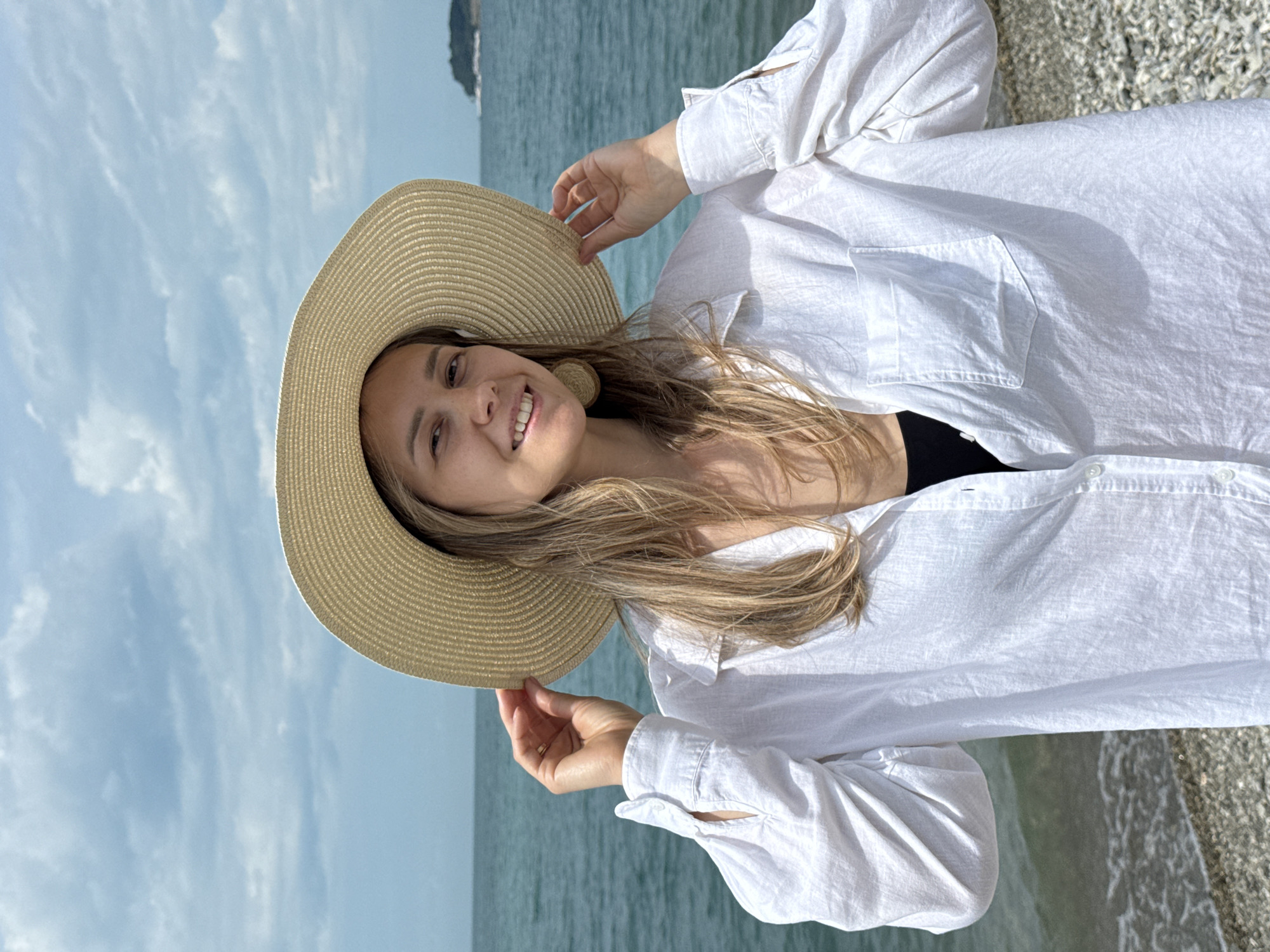 A woman stands on a pebble beach in Nebug, Russia, wearing a wide-brimmed straw hat and a loose white button-down shirt over a black top. She is smiling and holding the brim of her hat with her left hand. The ocean is visible behind her, with a small island or landmass in the distance. The sky is cloudy, with patches of blue. The woman has blonde hair and is wearing large, circular earrings. The lighting suggests it is daytime.