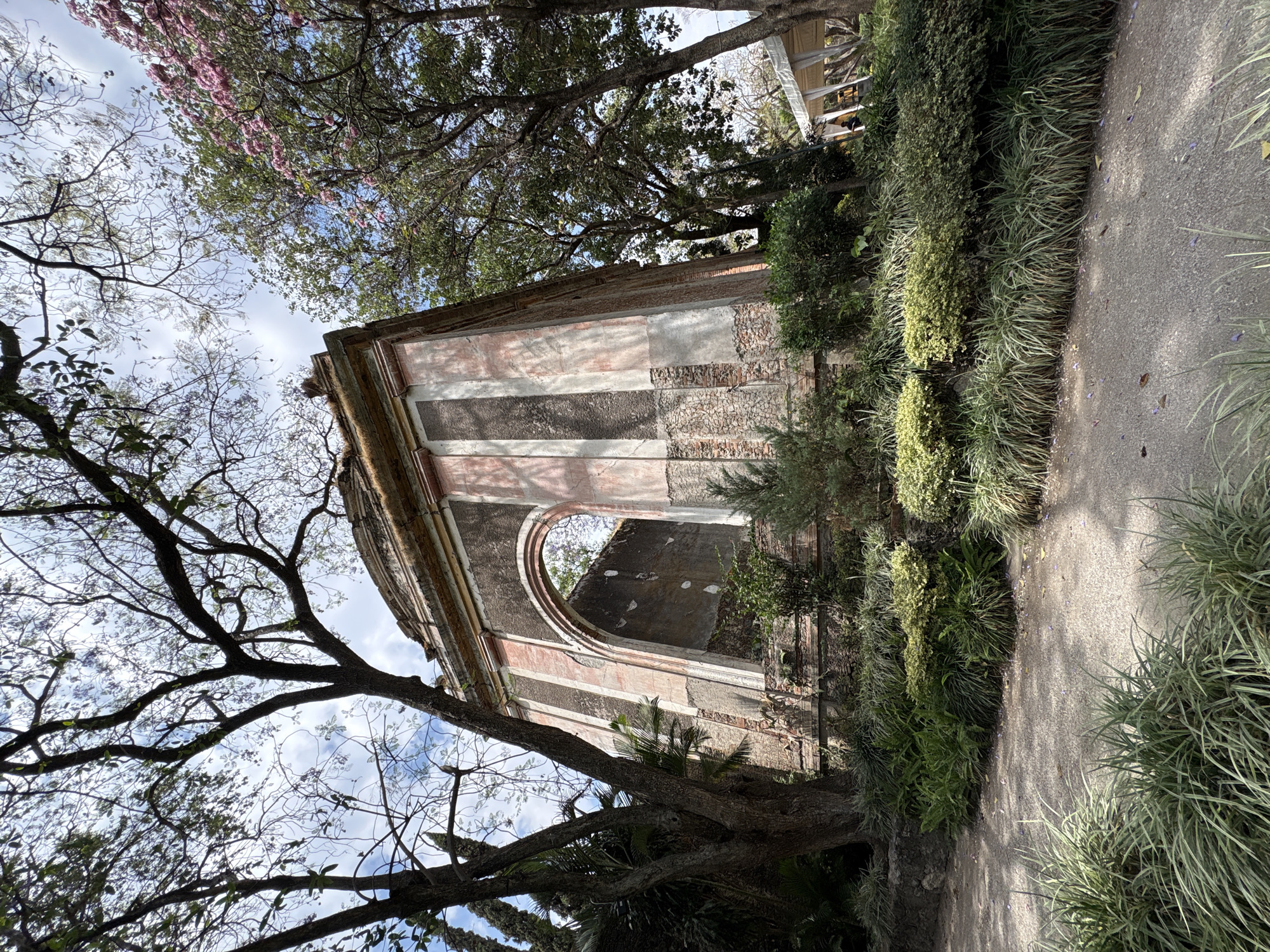 The image shows a ruined building with an arched opening, featuring pink and grey striped accents. The building is surrounded by lush vegetation, including trees with bare branches against a partly cloudy sky. A paved path borders the greenery. The scene appears to be located in a garden or park setting. This structure resembles the ruined building at Parque de las Esculturas in Progreso, Mexico.