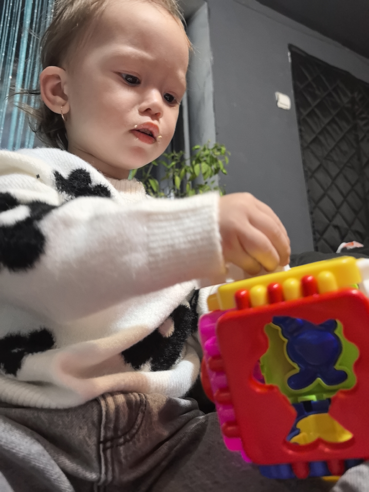The scene depicts a young child indoors, located in Noviy, Russia. The child, with light brown hair and a small gold hoop earring in the left ear, is seen from the upper torso, wearing a white sweater with black irregular patterns and grey pants. Small food particles are present on the child's mouth. The child's right hand is engaging with a multi-colored plastic toy, which is a shape sorter or similar item, featuring a red main section with a blue and yellow figure, a yellow top with red protrusions, and pink and blue sides. The background consists of a plain dark grey wall. To the left, vertical turquoise elements, possibly decorative strings or blinds, are partially visible. A small green potted plant is situated behind the child's left shoulder. To the right, a dark, quilted door is visible, above which a small white rectangular object is affixed to the wall.