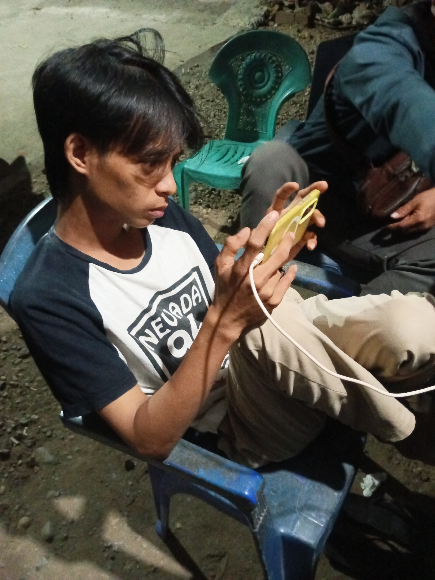 In Tumampua, Indonesia, a young man sits on a blue plastic chair, holding a yellow smartphone with a white charging cable plugged in. He is wearing a black and white raglan t-shirt with "NEVADA OL" printed on it. His pants are a light tan color. In the background, another person is partially visible, wearing a dark jacket and carrying a brown bag. A green plastic chair is also present in the background. The ground appears to be a mixture of dirt and gravel. The lighting suggests it is either dusk or nighttime.
