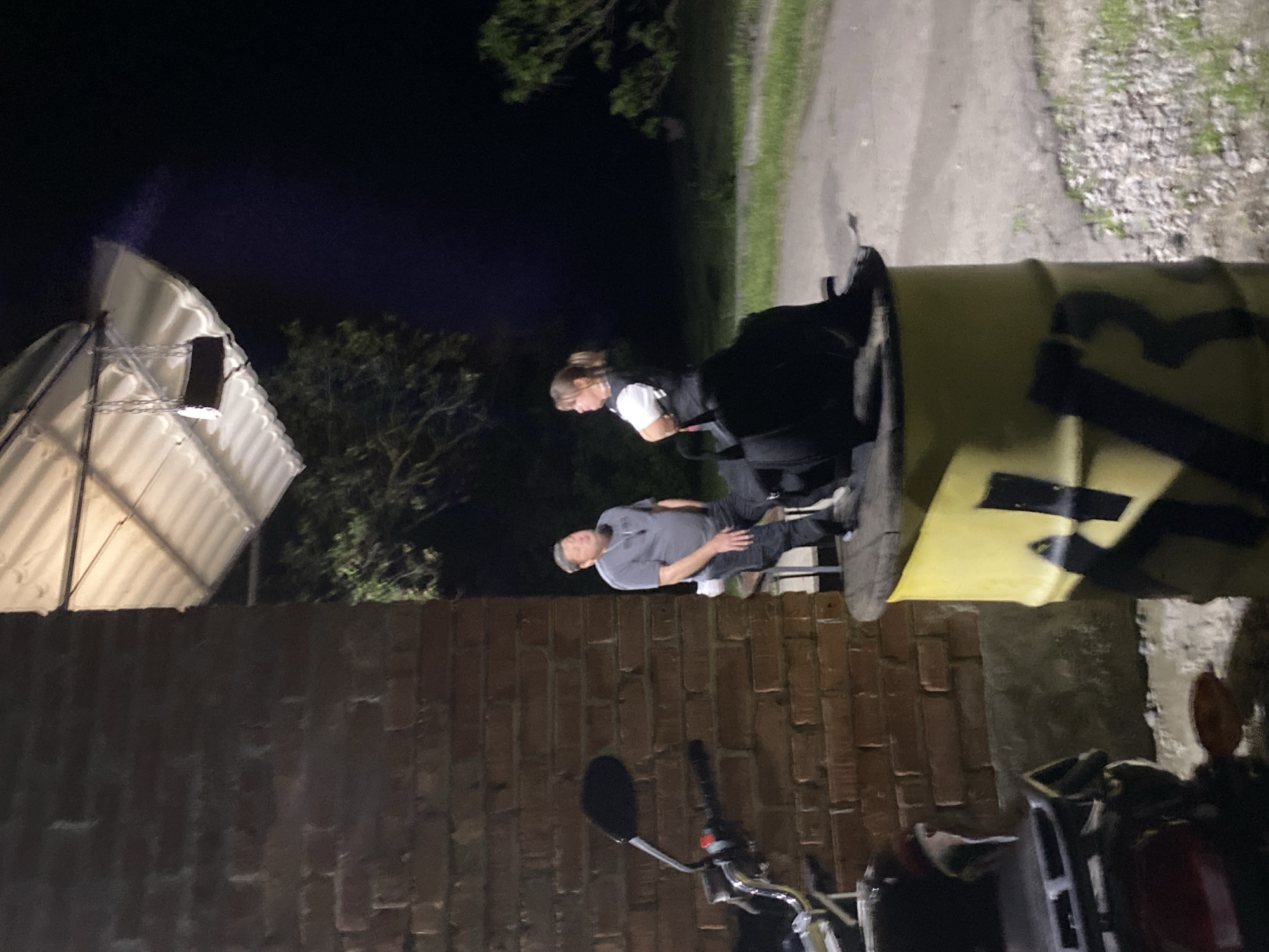 The image depicts a scene outdoors at night. A man in a gray shirt and dark pants is lying down. A woman wearing a white shirt and a dark vest is standing nearby. They appear to be next to a large yellow barrel or container with black markings. There is a brick wall and a motorcycle in the foreground. A white structure with a sloping roof is visible in the background, alongside trees. Based on the prompt, the location is Ustyvytsia, Ukraine, but without further details, specifics about the location cannot be stated.
