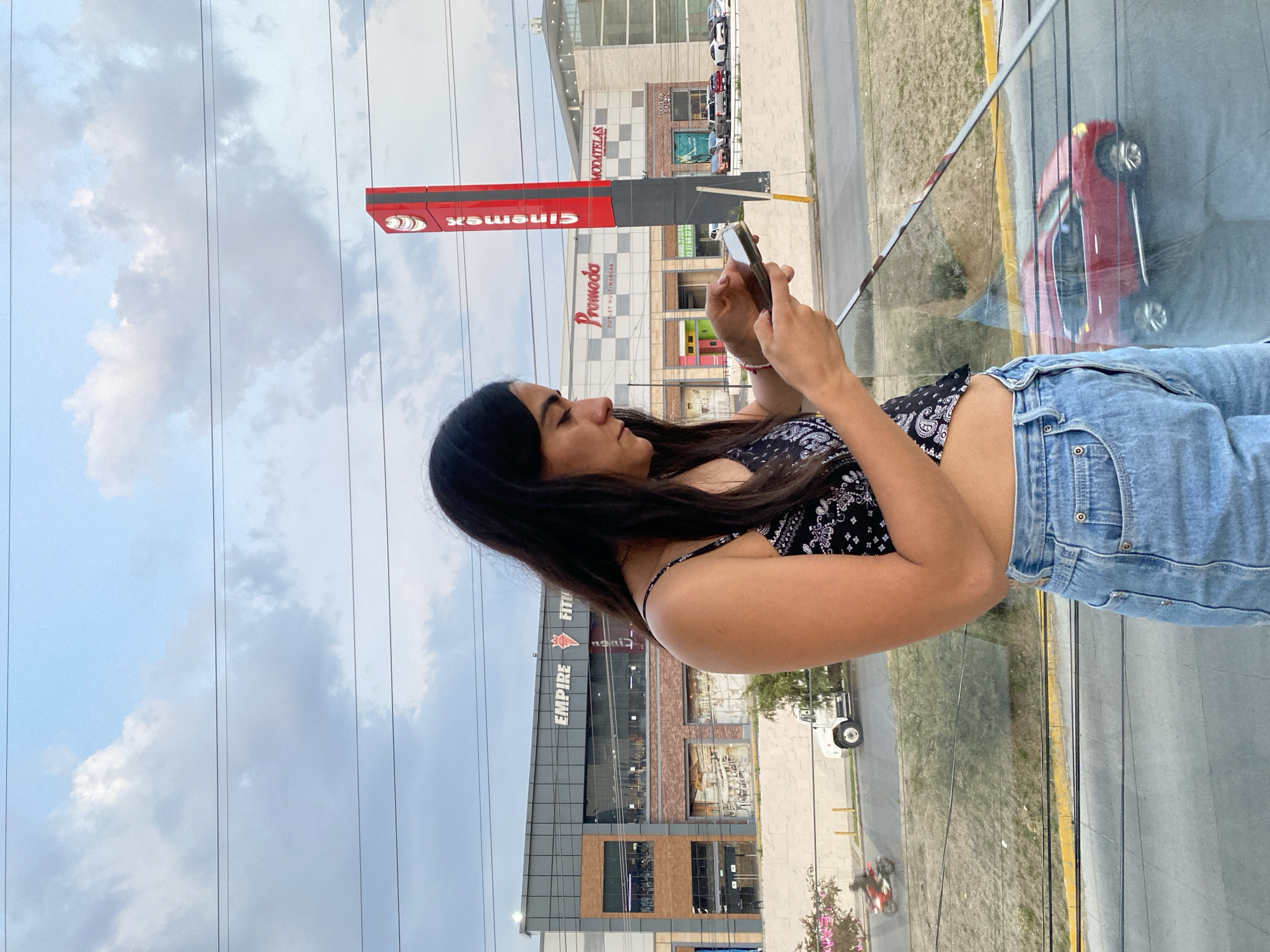 A young woman with long, dark hair is holding a smartphone. She is wearing a black and white patterned top and light-wash denim jeans. The background shows a city scene, including buildings and a street. Buildings visible include "Cinemex" and "Promoda Outlet Multimarcas." Also, a building with "EMPIRE FITNESS" written on it is shown. A red car is reflected on the glass pane. Overhead power lines are visible. The location is Puebla, Mexico, based on the brand names of the businesses.