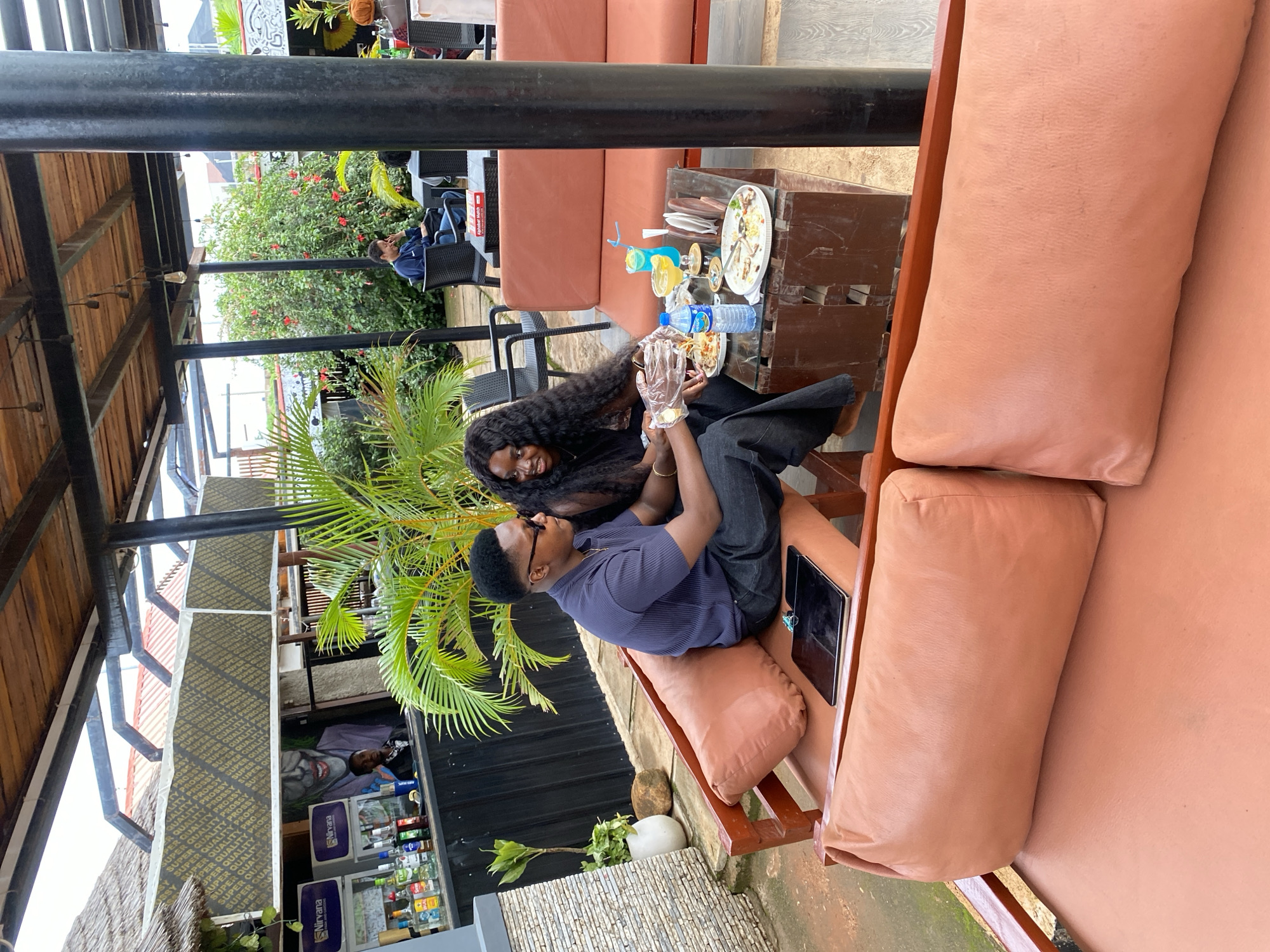The image depicts an outdoor seating area at a bar or restaurant in Gura Top, Nigeria. In the foreground, a couple is seated on a large, tan sofa. The man, wearing a navy blue ribbed polo shirt and dark pants, is seated on the left, looking to his right. The woman, with long dark curly hair and wearing a black top, is seated on the right, also looking towards the man. They are both holding plates of food. In front of them, on a low, dark brown wooden table with a glass top, are two blue cocktails, a bottle of water, and plates with leftover food. A tablet is visible on the sofa to the man's left.
Behind the couple, a bar area is visible with various bottles of alcohol displayed. A large mural of a person's face, possibly the Joker, is painted on the wall behind the bar. To the left of the bar, a potted plant stands near a textured stone wall. Lush green plants and trees are scattered throughout the outdoor area, with some red flowers visible on a bush in the background. Other patrons are seated at tables in the distance, and a black pillar supporting the overhead structure is prominent on the right side of the frame. The overhead structure features a wooden ceiling with exposed beams and hanging lights.