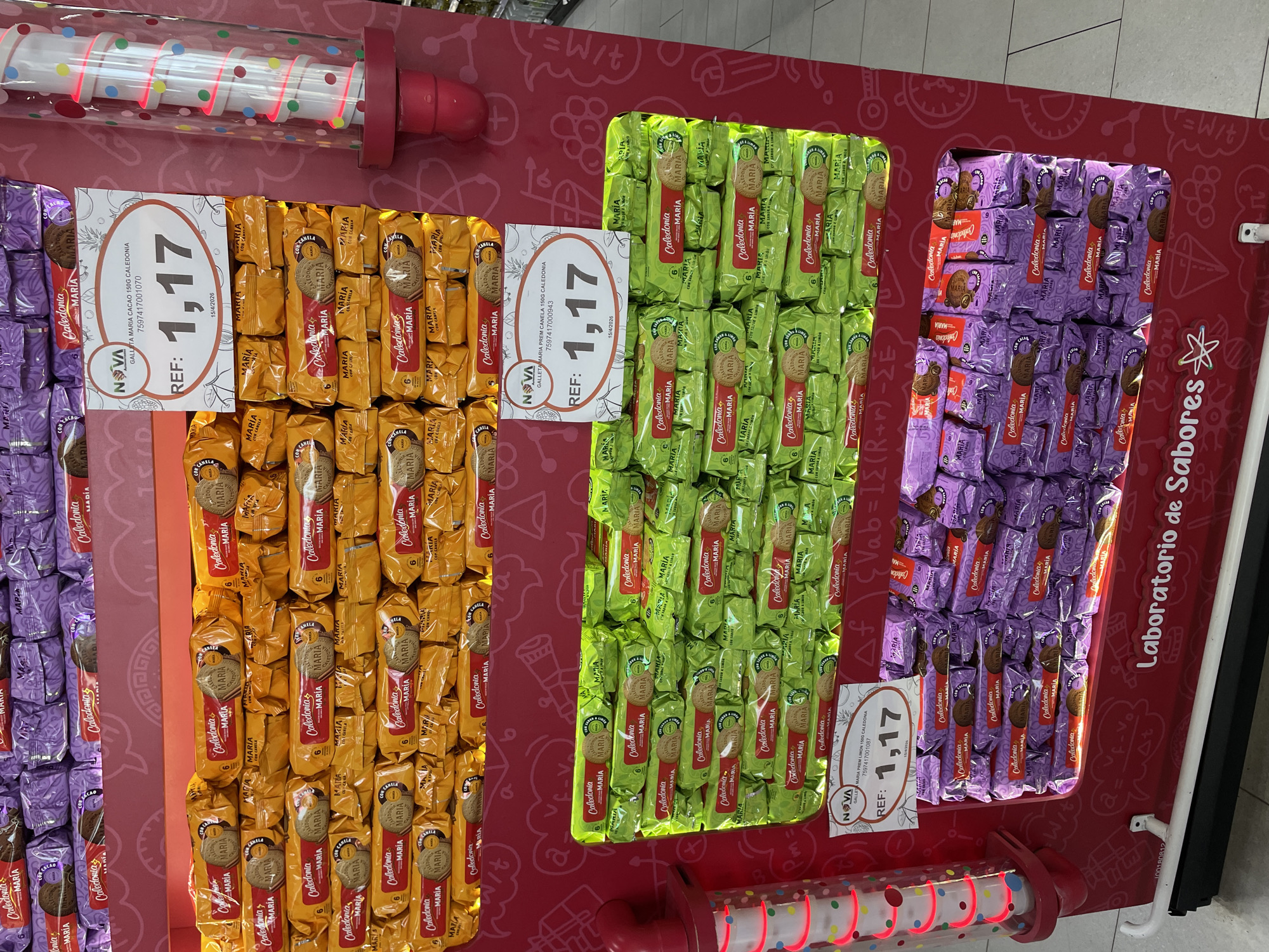 The image depicts a retail display of packaged cookies in a store, likely a supermarket or convenience store, in Porlamar, Venezuela. The display features multiple shelves stacked with cookies in brightly colored packaging, primarily yellow, green, and purple. Each package prominently displays the brand name "Caledonia" and "Maria" along with images of the cookies themselves.
Several price tags are visible, indicating a price of "1,17" for various cookie varieties. One price tag specifies "GALLETA MARIA CACAO 150G CALEDONIA" and another "GALLETA MARIA PREM CANELA 150G CALEDONIA". The overall environment suggests a well-stocked retail setting. The lighting is bright, typical of a commercial establishment. There are no people visible in the immediate foreground of the image. The background shows more shelving and possibly other merchandise, but it is out of focus.