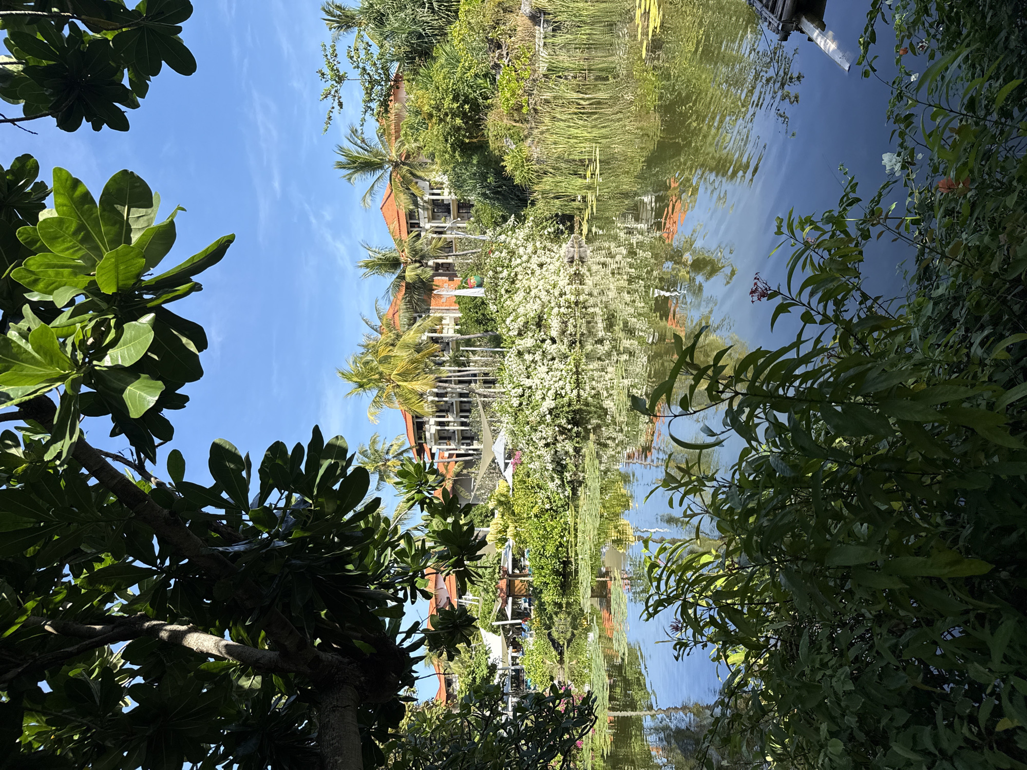 The image shows a tropical scene with lush vegetation and a building reflected in the water. The photo seems to have been taken from a low angle, with trees and foliage framing the view. In the background, there is a building with an orange tiled roof, possibly a resort or hotel, with palm trees surrounding it. The reflection of the building and trees can be seen in the water, suggesting that it is a lake or a pond. Some plants grow along the shore with white flowers. The blue sky is visible in the background.
The location is in Kuta Selatan, Indonesia, as indicated in the prompt.