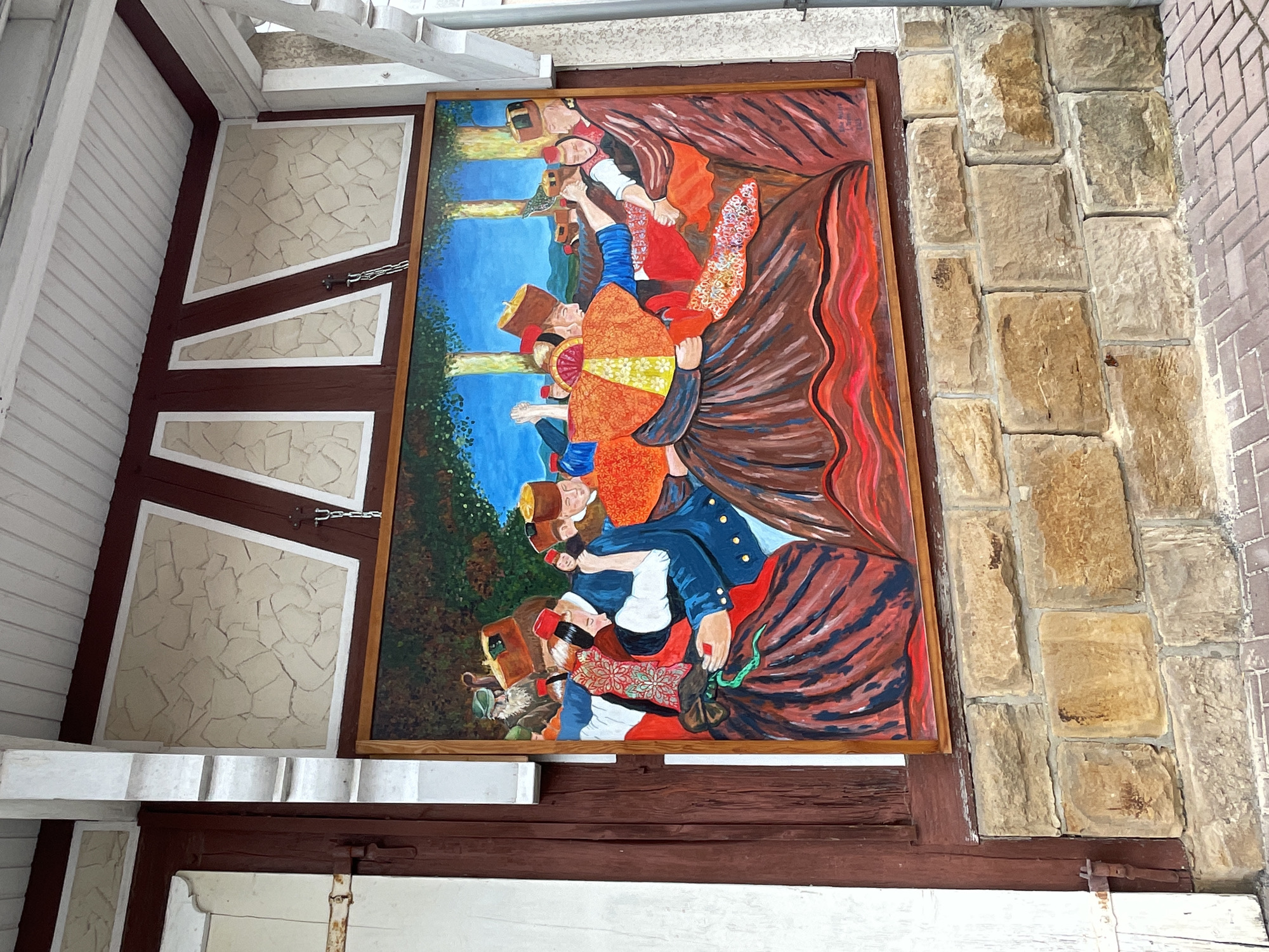 The image shows a large, framed mural depicting a scene of people in traditional attire dancing. The mural is mounted on a stone wall beneath a covered walkway with a wooden ceiling. The artwork features men and women dressed in brightly colored clothing, including hats and full skirts, with an overall folk art style. The background of the mural includes trees and a blue sky. The stone wall is constructed of irregularly shaped tan and brown stones. The walkway area is bordered by a white railing and has wooden beams supporting the roof. The area also features some architectural elements with a textured, cream-colored infill framed by dark red trim. A portion of a white wooden structure with a metal latch is visible on the left side of the image. The mural is located in the city of Willingshausen, Germany, known for its artist colony.