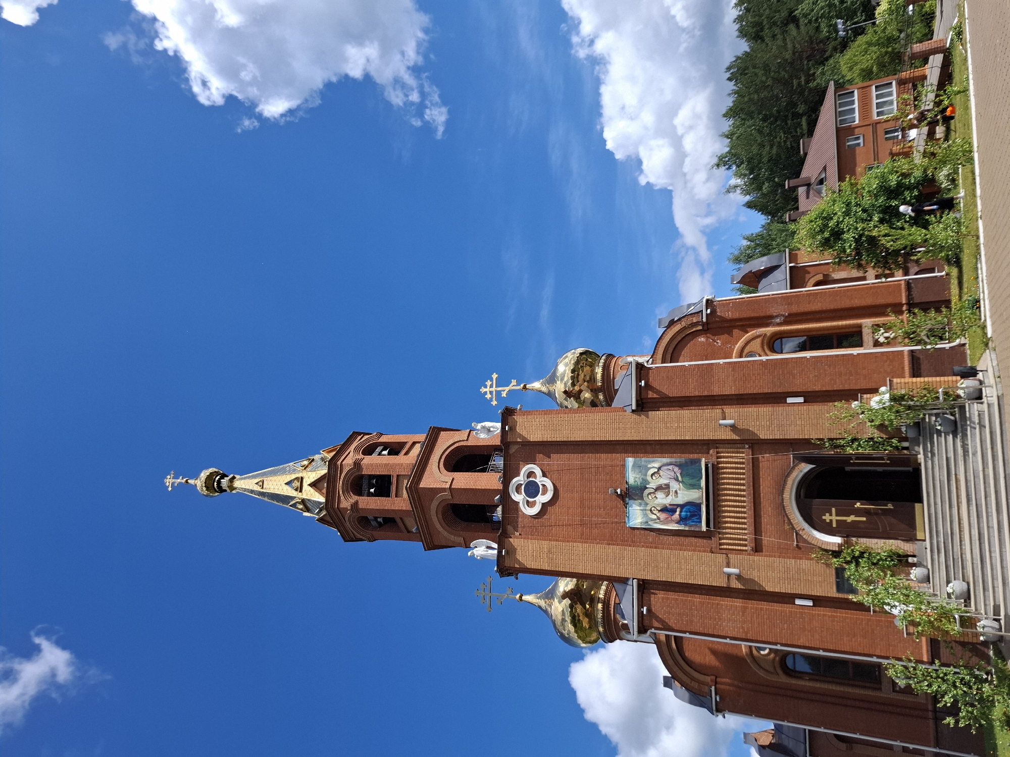 The image depicts the Church of the Holy New Martyrs and Confessors of Russia, located in Chaikovsky, Perm Krai, Russia. It is a brick building with golden domes and crosses. The church has a prominent bell tower with a golden spire. A religious painting is displayed on the exterior of the church. Stairs lead to the entrance. The sky is blue with some white clouds. Adjacent to the church is a building, and trees.