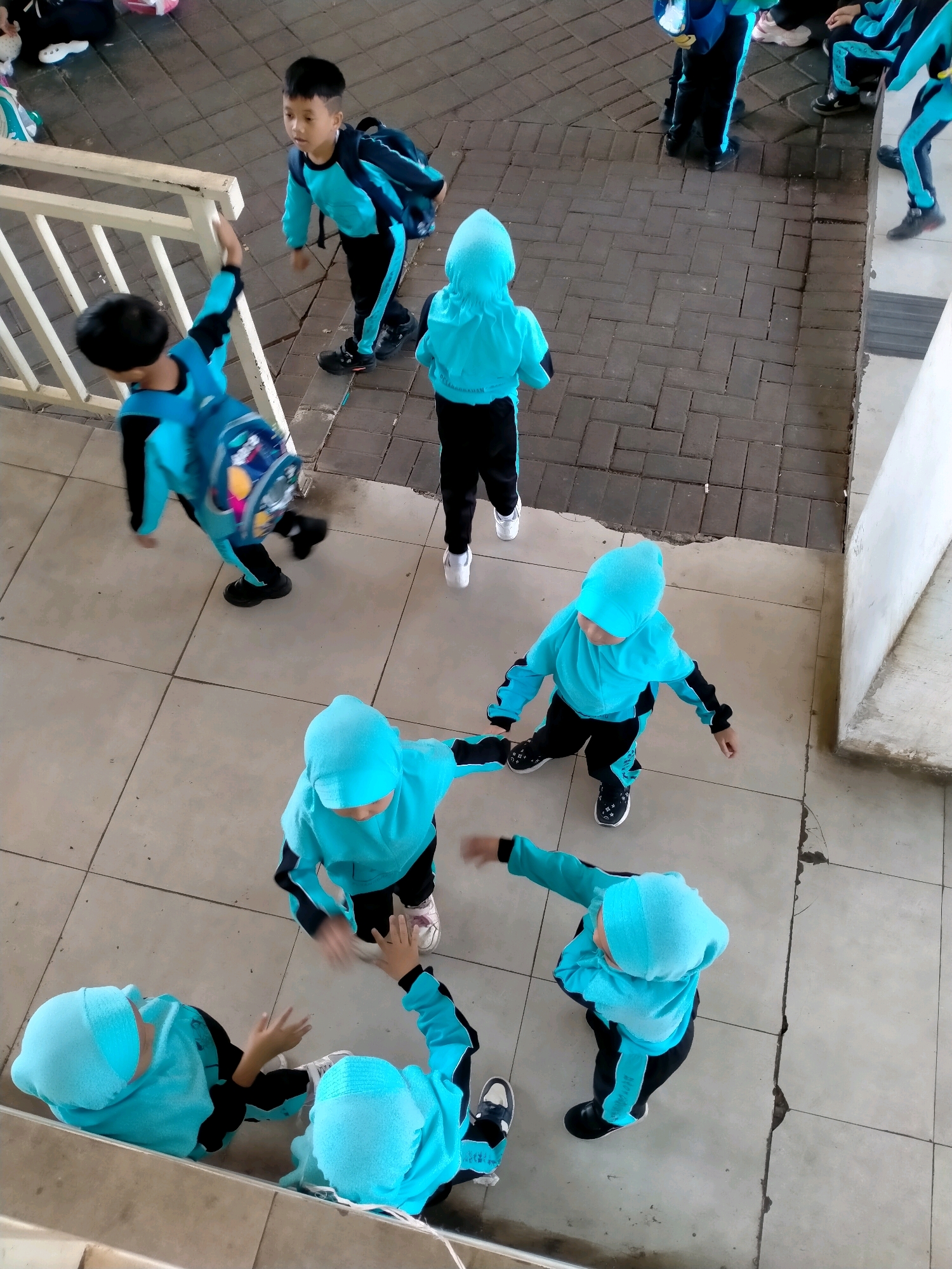 The overhead shot captures a group of children in turquoise school uniforms with black accents and hoods, some wearing matching headscarves, in what appears to be an outdoor educational facility in Batu, Indonesia. The children are distributed across a tiled floor and a paved area. In the background, two boys in similar uniforms are visible, one facing left and wearing a backpack, the other facing right. A white railing separates the tiled area from the paved background. In the foreground, a group of four children in turquoise uniforms are engaged in various activities. One child is walking, another is in a crouched position, and two others appear to be interacting, possibly playing or engaging in a lesson. The overall scene suggests a school environment, likely a playground or an outdoor learning space, within the city of Batu, known for its educational institutions and cooler climate.