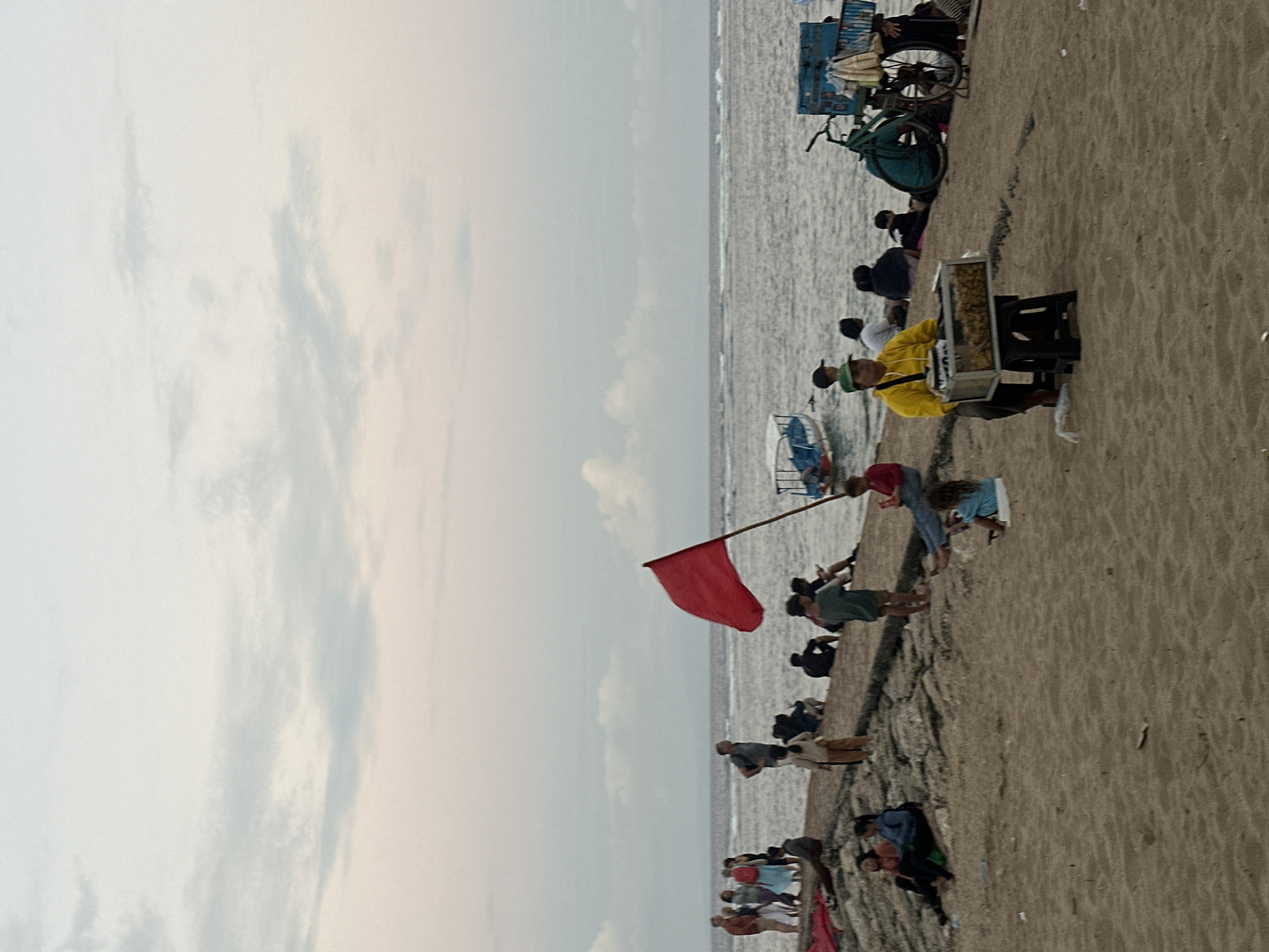 In Denpasar, Indonesia, a group of people are gathered on a sandy beach near a stone jetty. The sky is overcast with soft clouds. A red flag waves from a pole on the jetty, signaling potentially hazardous water conditions. Several individuals are seated or standing on the jetty, looking out at the ocean, while others are scattered on the sand. A vendor in a yellow jacket sits on a stool next to a food cart displaying fried items, possibly for sale to beachgoers. In the background, a small blue and white boat is visible in the water, and a bicycle with a cart attached is parked on the sand. The overall scene depicts a casual gathering at the beach, with elements of local commerce and a clear warning about the sea.