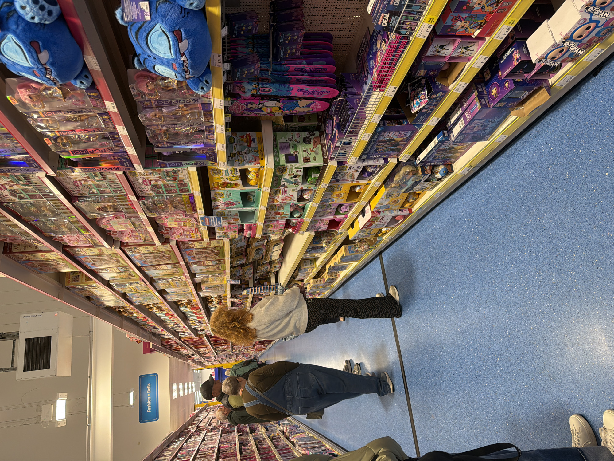 The image shows an aisle in a retail store, likely a toy section, located in Wrexham, United Kingdom. A blue floor and shelves stocked with various toys and games define the scene. Customers are browsing the merchandise, including adults and children. "Fashion Dolls" signage is visible. Shelves are filled with items like "K-POP idol" dolls, puzzles (Ravensburger), and games such as Monopoly Deal. Price tags are displayed on the shelves. A Powrmatic HVAC unit is mounted on the wall.