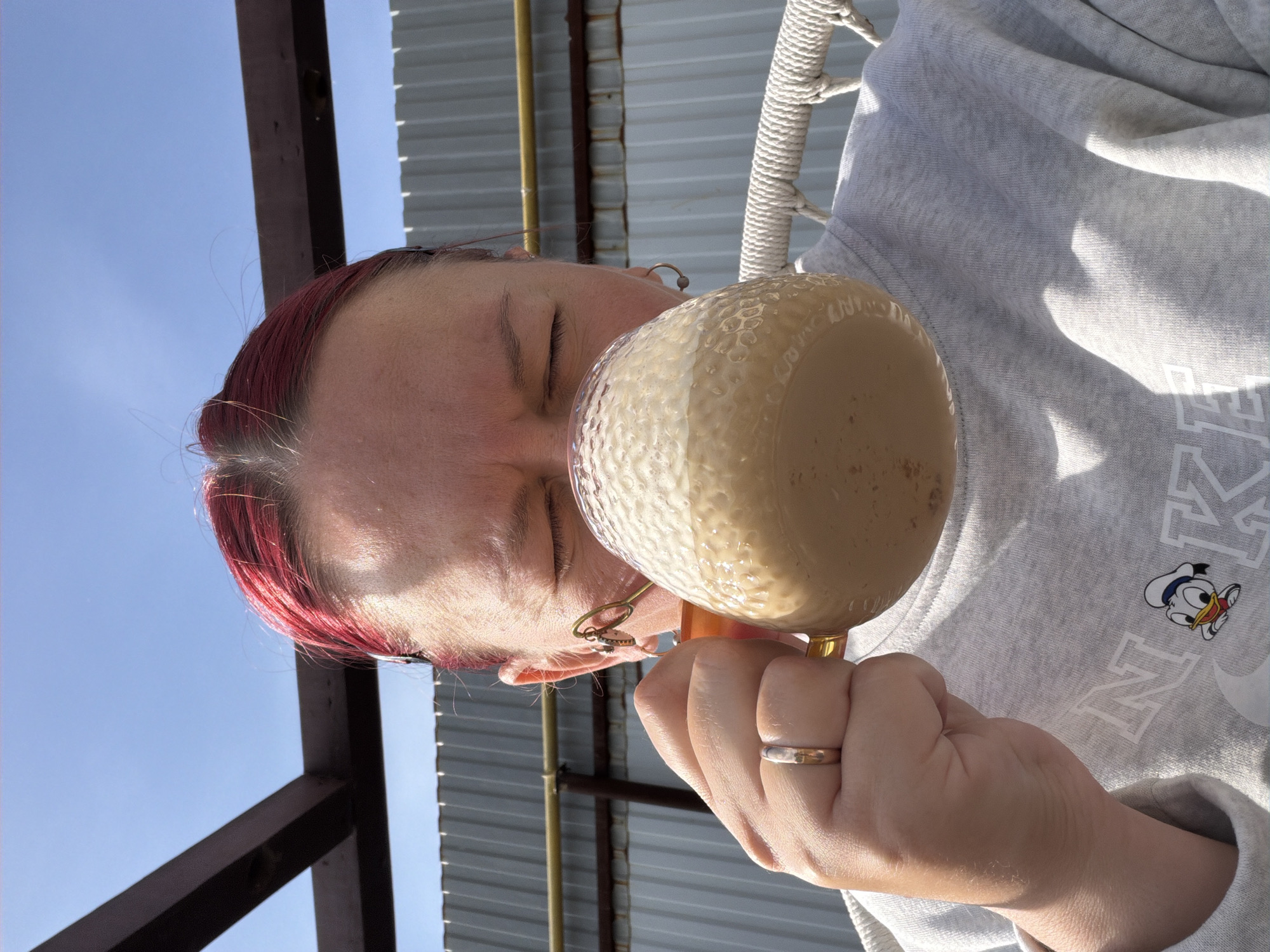 A woman with red hair, wearing a light gray sweatshirt with a Donald Duck cartoon, holds a large textured beige mug filled with a light brown liquid against her face, obscuring her mouth. Her eyes are closed. She wears gold earrings and a wedding band on her left hand. The background includes blue sky, brown wooden beams, and a corrugated metal roof. The scene appears to be outdoors, possibly on a patio or porch. No information is available to identify the specific location as Chaikovsky, Russia.