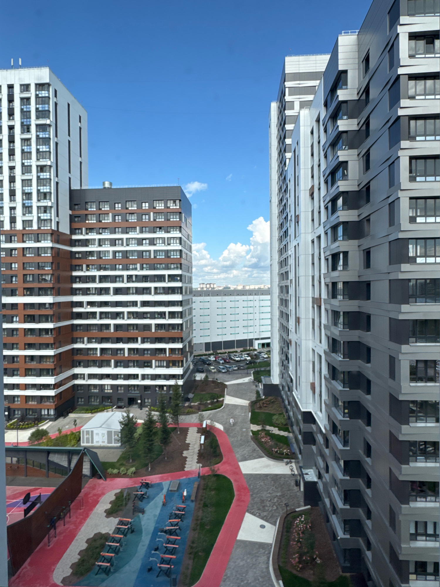 The image captures a modern urban landscape with multiple high-rise apartment buildings under a clear blue sky with scattered white clouds. The buildings display contemporary architectural designs with varied facades. In the foreground, a courtyard area is visible, featuring a children's playground with colorful surfaces and exercise equipment. The ground is paved with a mosaic of stone and red pathways, interspersed with greenery and trees. A white, tent-like structure is situated in the middle ground, possibly a community space or temporary facility. A parking lot with numerous cars is present further back, adjacent to a large, low-rise building. The scene suggests a residential complex, likely in a developed area of a city. The location is identified as Novosibirsk, Russia, specifically the "Europeisky Bereg" residential complex.