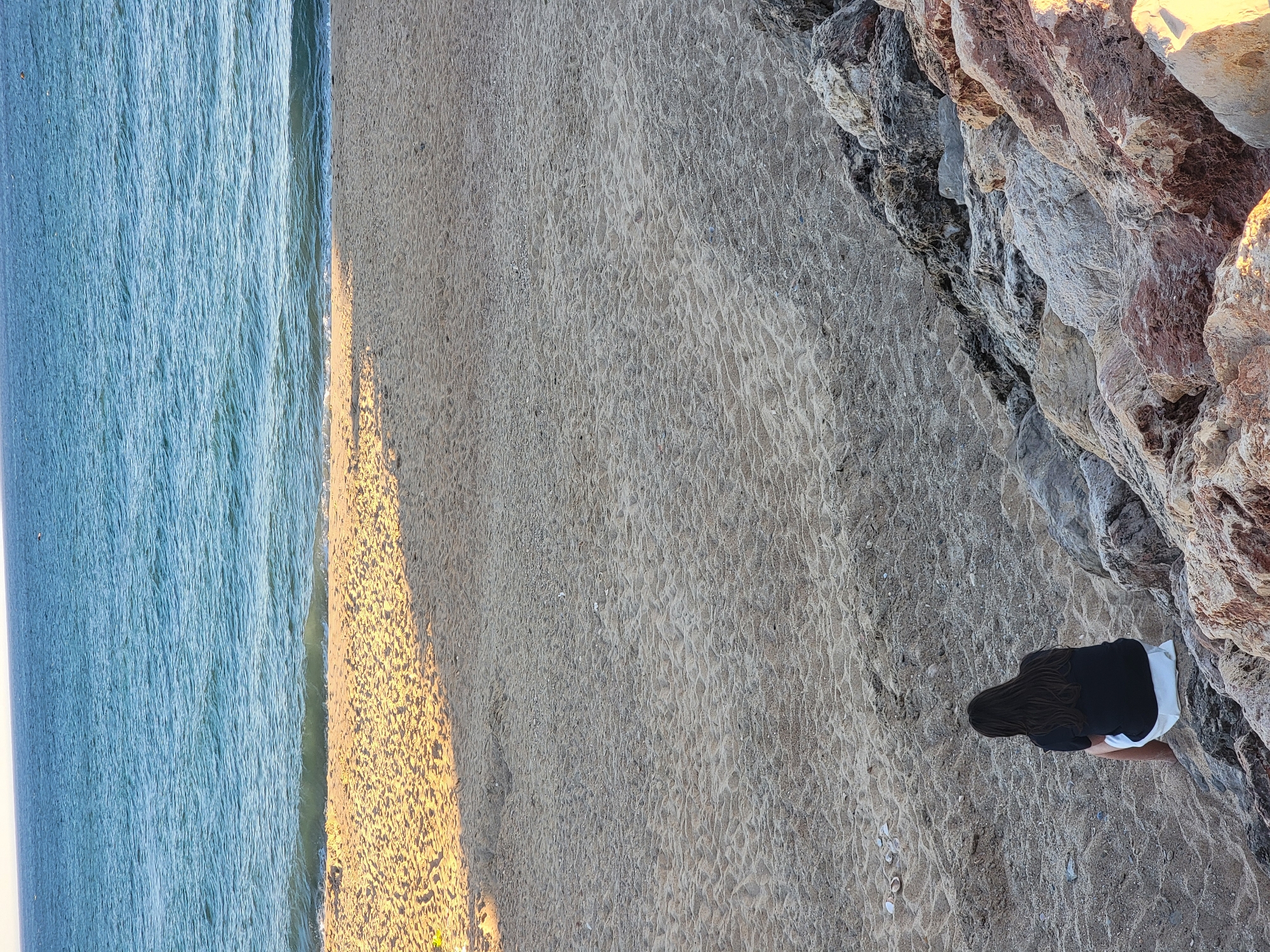 The image captures a serene beach scene in Quarteira, Portugal. In the foreground, a person with dark hair, wearing a black t-shirt and white shorts, sits facing the ocean. They are positioned on rocky terrain that transitions into a sandy beach. The sand displays textured patterns, indicative of wind or water movement, and is dotted with small stones and shells.
The middle ground is dominated by the beach, with a clear demarcation between the dry sand and the wet sand where gentle waves are breaking. The water is a calm, deep blue, reflecting the clear sky above. The horizon line is sharp, separating the sea from the pale sky. Sunlight casts a long shadow across the sand, suggesting it is late afternoon or early morning. The overall impression is one of tranquility and natural beauty, characteristic of the coastal landscape of Quarteira.