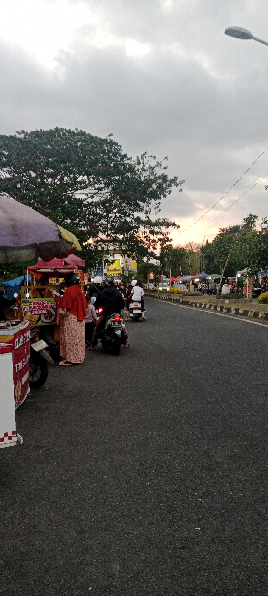This is a vertical, medium shot photograph taken at dusk on a street in Ngemplak, Indonesia. The scene depicts a roadside market with several food vendors and people on motorcycles. In the foreground, a food cart is visible on the left, displaying signage that reads "CHUNG GORE & TELUR BALUNG." Next to it, a woman in a patterned dress and red headscarf stands beside a food stall with an umbrella. A motorcycle is parked in front of the stall. Several other motorcycles are on the street, some carrying passengers, all moving away from the viewer and towards the right side of the frame. The road is asphalt, bordered by a curb and sidewalk where more stalls are set up under colorful umbrellas. Trees line the sidewalk, and the sky is overcast with a hint of sunset colors peeking through. A street lamp is visible in the upper right corner. The overall atmosphere is that of a busy evening street market.