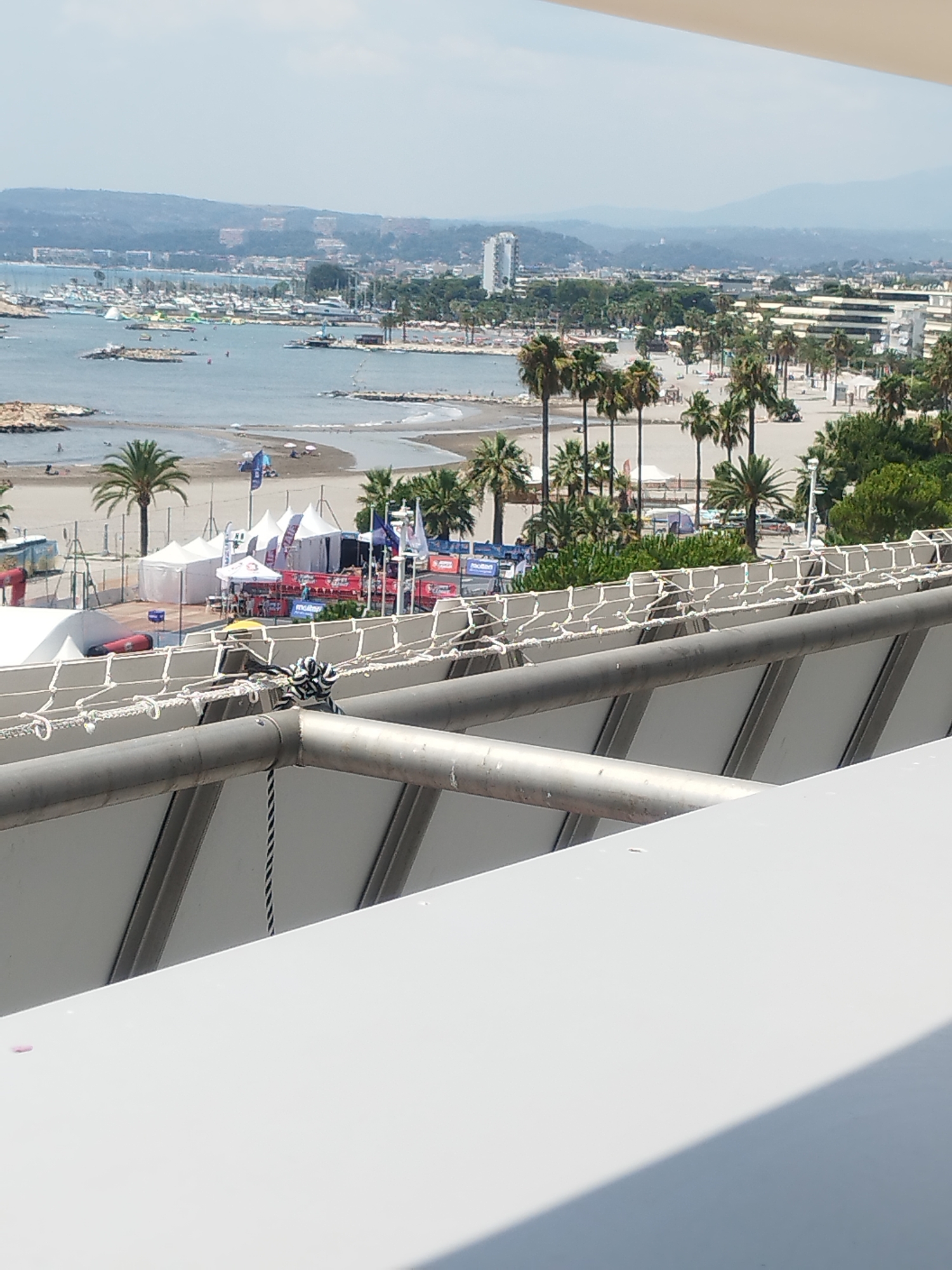 The image shows a view from an elevated position looking out over a bay and a coastal city. In the foreground, a white balcony railing with metal supports is visible, with a rope secured to the railing. Beyond the railing, the scene unfolds to reveal a busy harbor filled with numerous boats. The water is a calm, light blue, and several small islands or rocky outcrops are scattered within the bay.
The shoreline features a sandy beach with palm trees and beach umbrellas. Several white tents and structures, possibly for an event or market, are set up on the sand. In the background, the city of Saint-Laurent-du-Var, France, stretches along the coast, characterized by a mix of modern buildings and older structures, backed by rolling hills. The sky is clear and bright, suggesting a sunny day. There are no distinct individuals or specific actions clearly discernible in the image, but the presence of boats and beachgoers implies a recreational or tourist area.