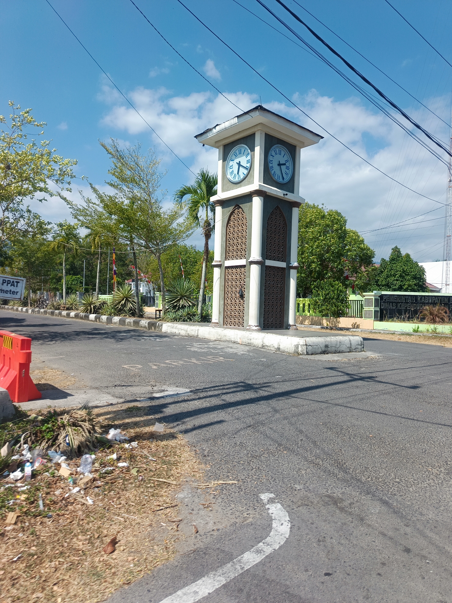 In Suwawa, Indonesia, a clock tower stands prominently on the side of a paved road under a clear blue sky with scattered clouds. The clock tower is square with a peaked roof and has two visible clock faces, each displaying Roman numerals and hands indicating the time. The tower is adorned with decorative lattice work. To the left of the clock tower, a sign with "PPAT meter" is visible. A bright orange traffic barrier is positioned on the roadside in the foreground, adjacent to a patch of dry grass and scattered litter. Beyond the clock tower, a green fence encloses an area with various trees and some buildings, including a sign that reads "PEMERINTAH KABUPATEN BO..." and "DINAS KEPENDUD..." suggesting a government office. Overhead, power lines stretch across the sky. The road surface shows tire marks and a white painted line.