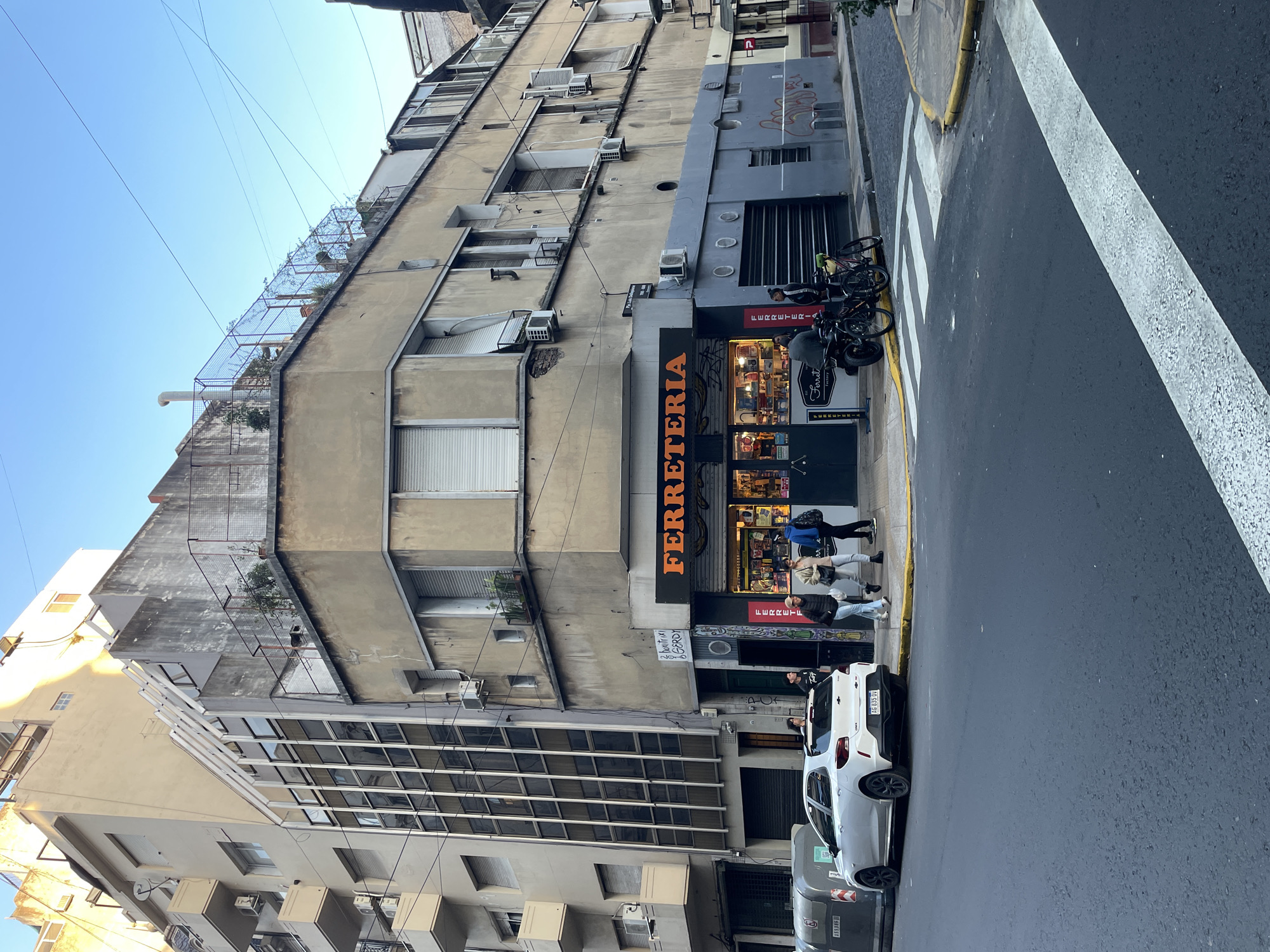 The image shows a street in Buenos Aires, Argentina. A multi-story building is on the left, housing a "Ferreteria" (hardware store) on the ground floor. Several people are near the entrance of the store, and bicycles are parked nearby. A white car, license plate AG 635 QV, is parked on the street in front of the store. The street has a crosswalk marked with white stripes. The sky is blue and clear. The building appears to be aging, with numerous air conditioning units attached to the façade. The building also features an enclosed rooftop area.