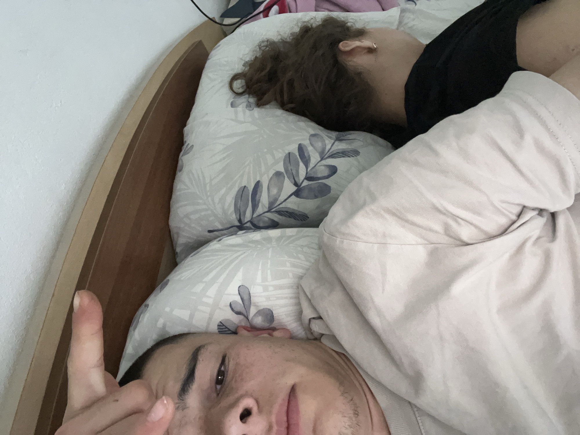 The image shows two people lying in a bed with a wooden headboard. One person is facing the camera with a finger raised. The other person is facing away from the camera with curly brown hair and is wearing a black shirt. They are lying on pillows with a leaf pattern and covered with a light-colored blanket. The environment appears to be indoors in a bedroom. No information in the image or associated hints indicates the location in the city of Karavelovo, Bulgaria.