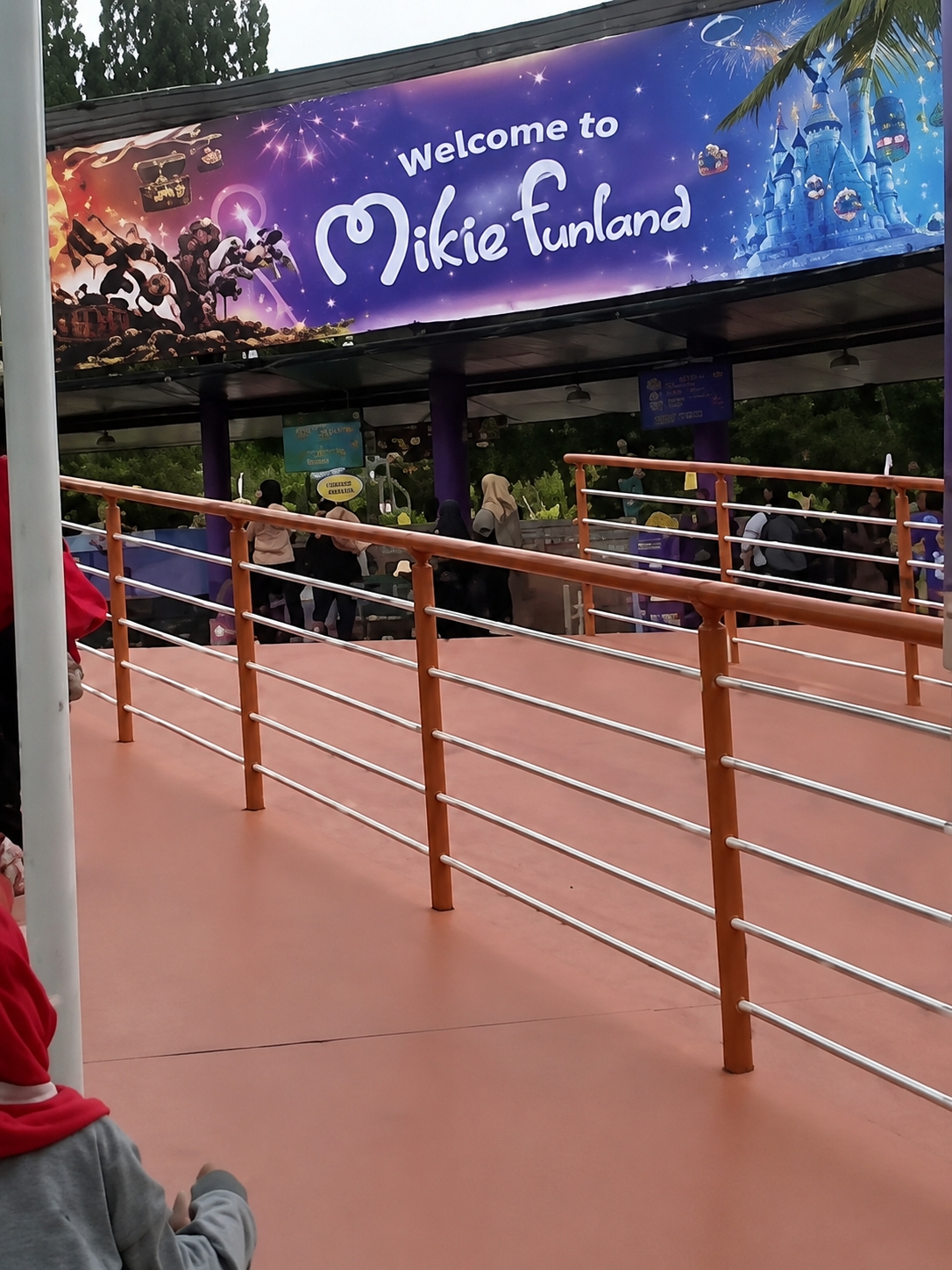 The image shows the entrance to Mikie Funland, an amusement park located in Berastagi, Indonesia. A large, colorful banner above the entrance reads "Welcome to Mikie funland" with an illustration of a blue castle and fireworks. Several people are visible in the background, queuing under a covered walkway. In the foreground, an orange railing separates the walkway from an orange-colored ground. A child wearing a red hooded jacket is partially visible in the lower left corner of the image, facing away from the camera. The environment appears to be outdoors, with trees visible in the background. The sky is overcast.