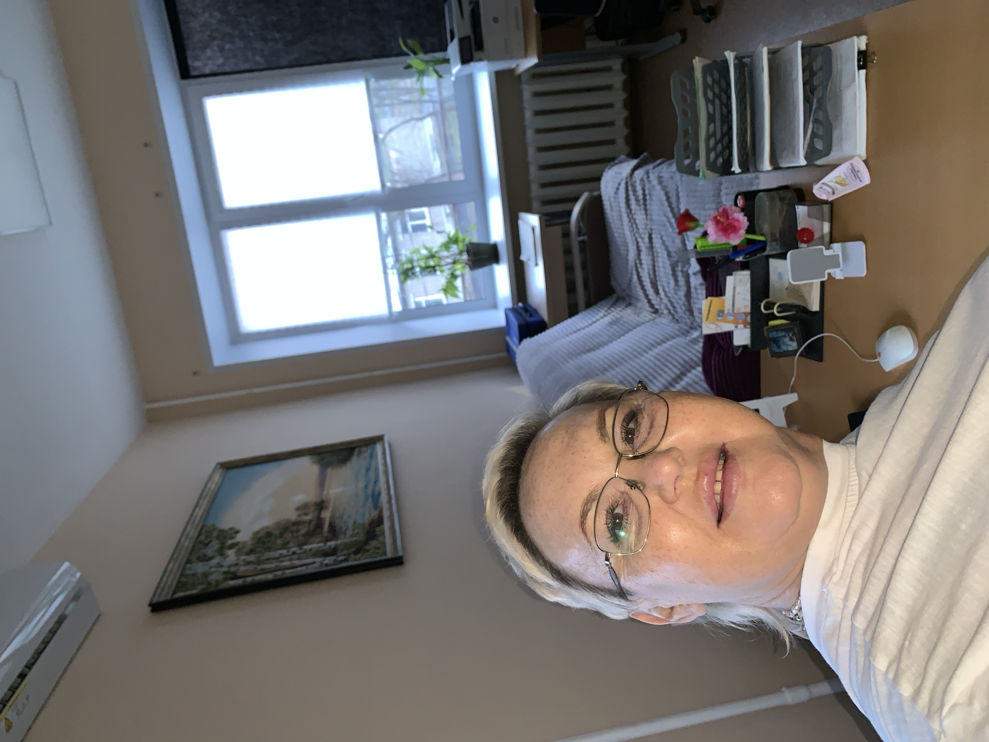 The image captures a selfie of a woman taken indoors, likely in an office or study in Chaikovsky, Russia. The woman, who appears to be middle-aged or older with light blonde hair and glasses, looks directly at the camera with a gentle smile. She is wearing a white top and a pearl necklace.
The background reveals a well-lit room, primarily from a large window on the far wall. Through the window, an urban or residential scene is visible with other buildings and bare trees, indicating a daytime setting. Below the window, a radiator is present. Two potted green plants sit on the windowsill, adding a touch of nature to the room.
To the right of the window, a desk or table holds various items including organized files in a rack, papers, a white computer mouse, and a small red flower (possibly a carnation or hibiscus) in a small vase. A beige tube of lotion with the partially visible text "SKIN CARE" and "PARIS" is also on the desk. Adjacent to the desk is a bed or sofa covered with a striped blanket. On the left wall, a framed landscape painting hangs above an air conditioning unit. The walls of the room are a light, neutral color. The overall impression is that of a comfortable and functional personal space.