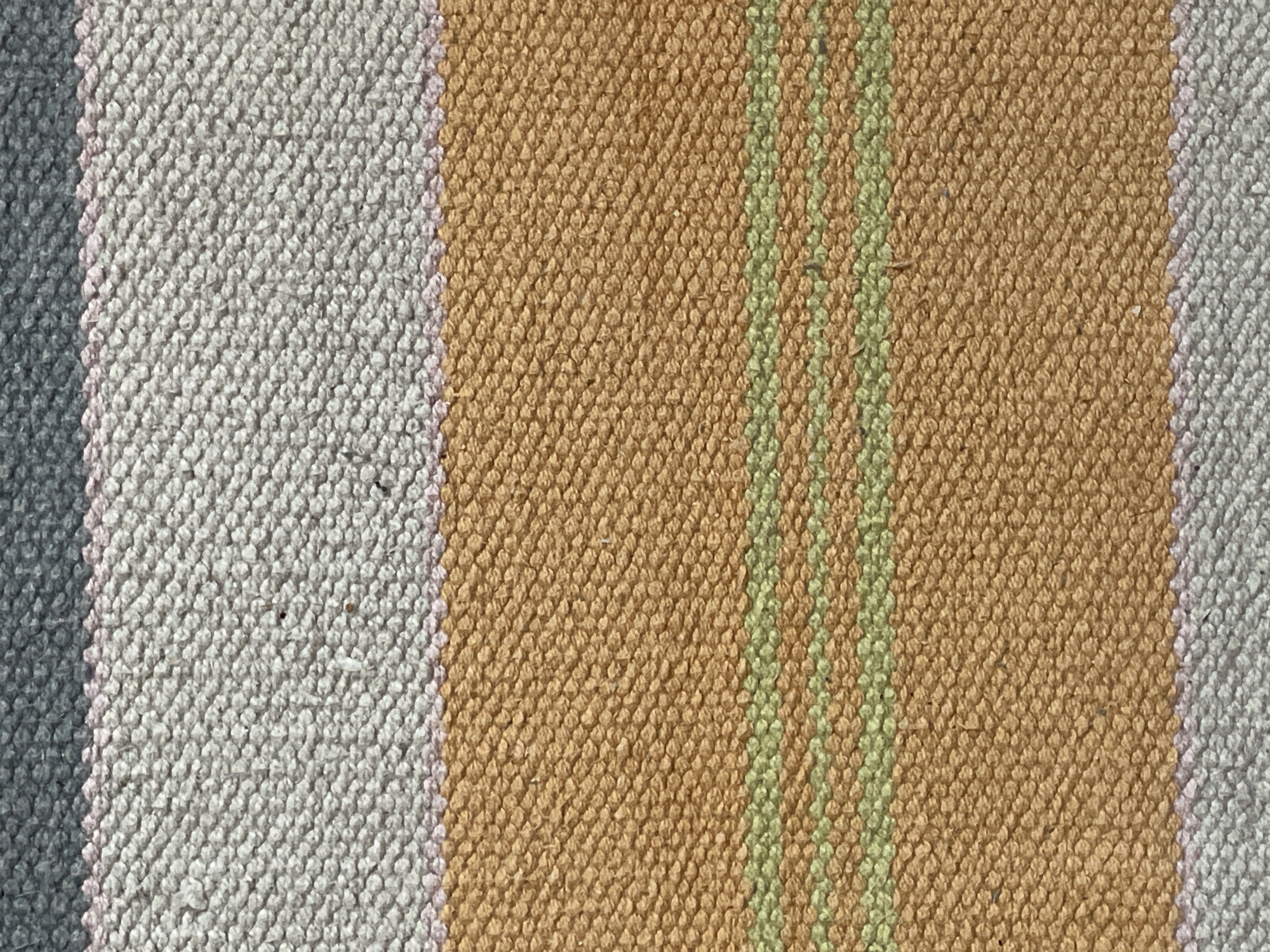 The image shows a close-up view of a textured fabric, likely a rug or tapestry. The fabric is divided into horizontal bands of color. From top to bottom, the bands are: dark gray, light pink, light gray, a wide band of orange-brown with two thin green stripes, and another band of light gray. The texture of the fabric is rough and woven, with visible individual threads. There are no people, objects, or environmental details present that would indicate a specific location in a city.