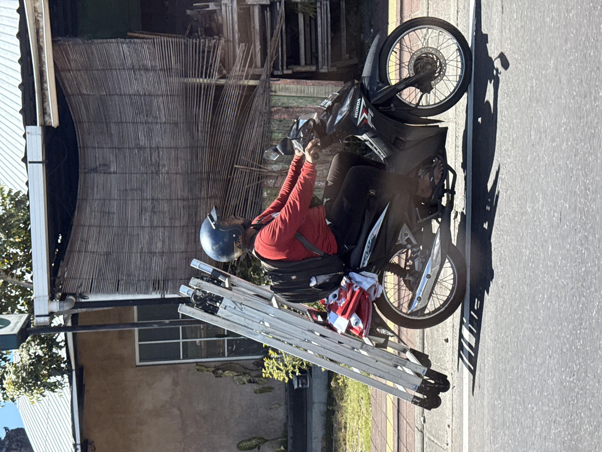 A person is seen riding a black Honda scooter, transporting a bundle of at least four large, silver folding ladders, and a red bag or item. The scooter is on a paved road with a white dividing line, next to a sidewalk with light-colored paving stones. The person, wearing a red long-sleeved shirt, black pants, a dark blue helmet, and sandals, is focused on driving. The ladders are securely strapped to the side of the scooter, extending significantly forward and backward.
The scene is brightly lit by the sun, casting long, distinct shadows of the scooter and its cargo onto the road, indicating it is likely morning or late afternoon. In the background, there is a building with a beige-colored wall and a white-framed window. Above the building, a large, dark-colored woven bamboo screen or shutter is visible, along with parts of a white gutter system. Lush green foliage and trees are also present on the left side of the frame. The scooter prominently displays "HONDA" on its side panel and "EVO" or "EVOL" on the rear seat. The person's helmet appears to have faint branding as well. This street scene is characteristic of daily life in places like Kuta, Indonesia, where motorcycles are a common mode of transport for both people and goods.