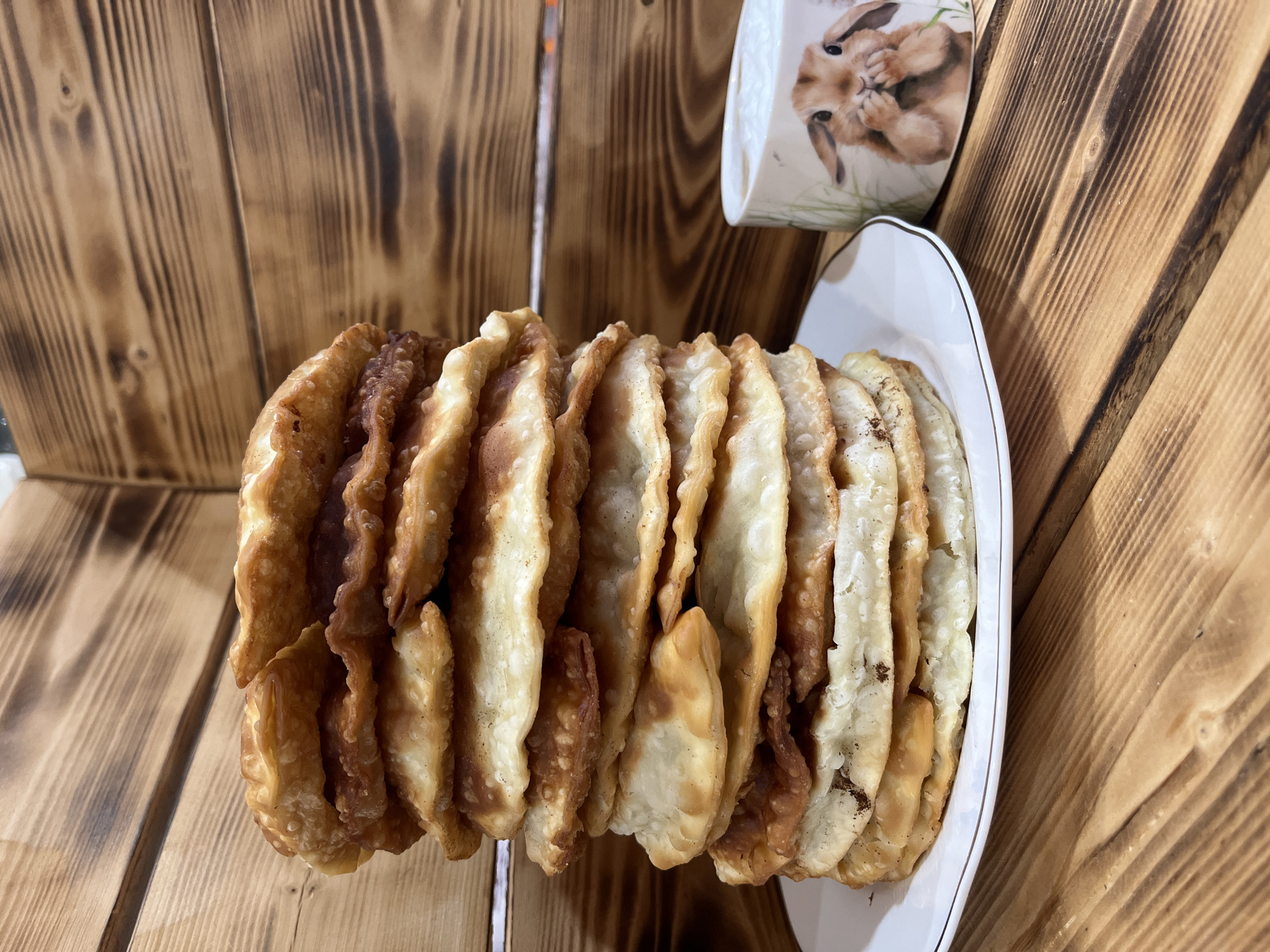 The image shows a stack of fried pastries on a white plate with a gold trim. Behind the plate is a mug with a bunny illustration and a backdrop of wooden planks. The location is Chaikovsky, Russia.