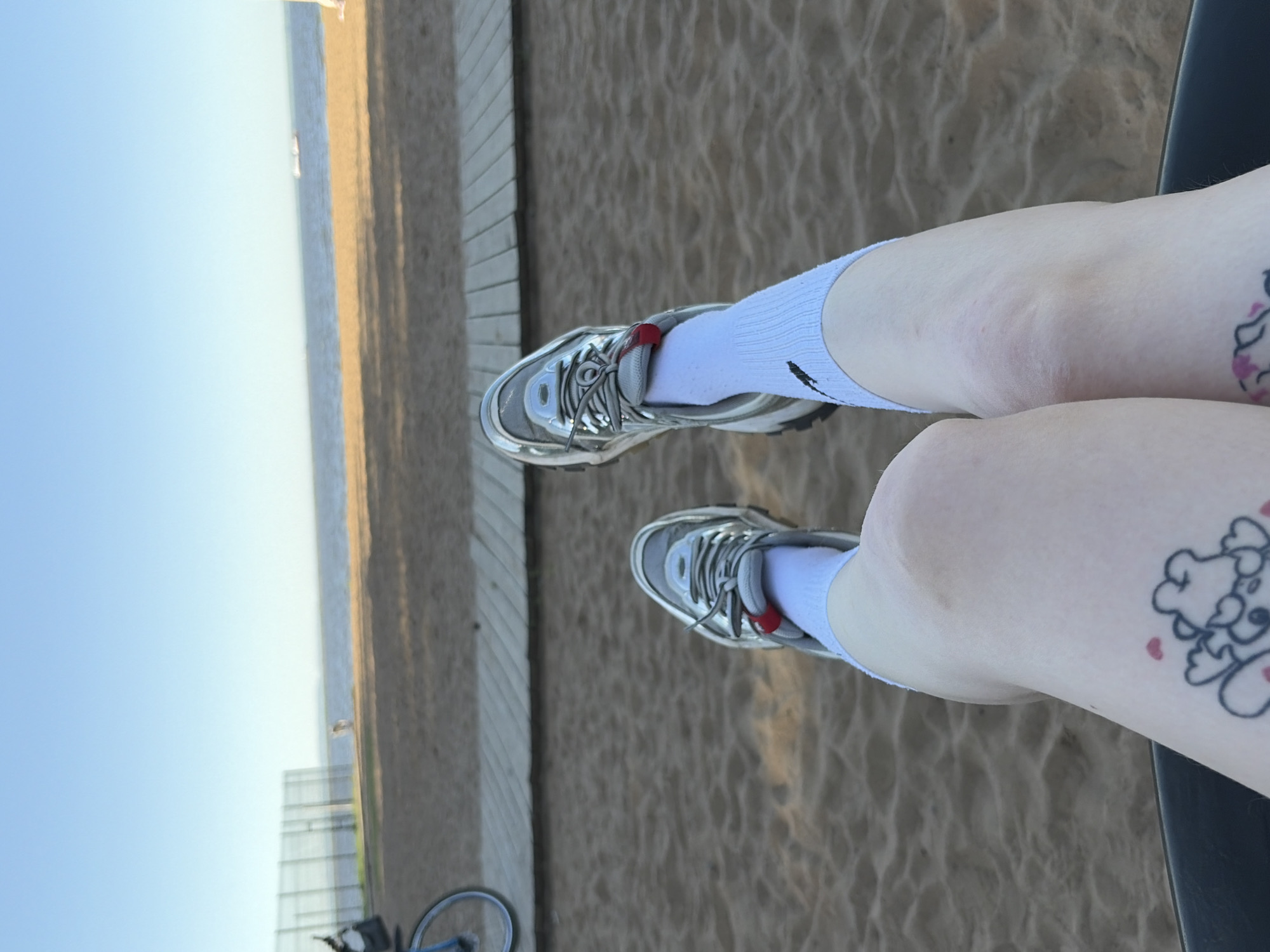 A low-angle, rotated shot features a person sitting on a black chair with legs crossed and knees visible, wearing white socks and silver sneakers. Each knee bears a black-ink tattoo of a cloud design with pink heart accents.
The background reveals a sandy beach extending towards a body of water under a light-blue sky. Adjacent to the beach is a wooden boardwalk. The image is taken outdoors during daylight. To the left, there are hints of buildings and a bicycle. The location is a beach environment, with no city identified.