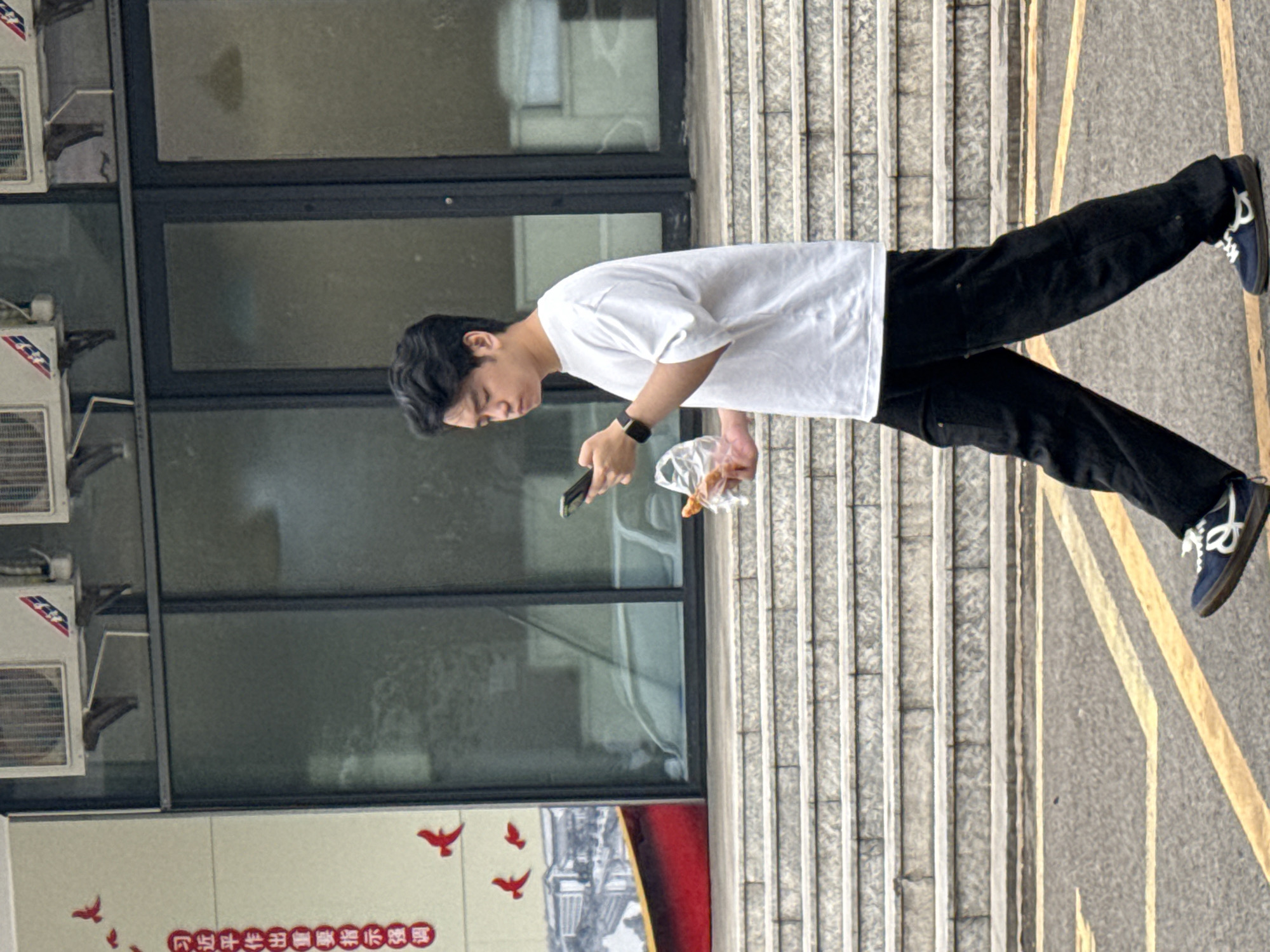 In a medium shot taken outdoors in Jiangjin, China, a young East Asian male, appearing to be in his late teens or early twenties, walks across a paved area with yellow lines. He is facing left and looking down at his smartphone, which he holds in his right hand. His left hand clutches a plastic bag containing a pastry. He is wearing a loose-fitting white t-shirt, black pants, and dark blue low-top sneakers with white laces and accents.
Behind him, a wide set of concrete stairs with visible stone risers leads up to a building. The building features large glass windows and two air conditioning units mounted above the windows. To the left of the building, a wall displays vertical red Chinese characters and several red bird decals. The overall lighting suggests an overcast or diffused daylight. The camera is positioned at a low angle, slightly below the subject's eye level.