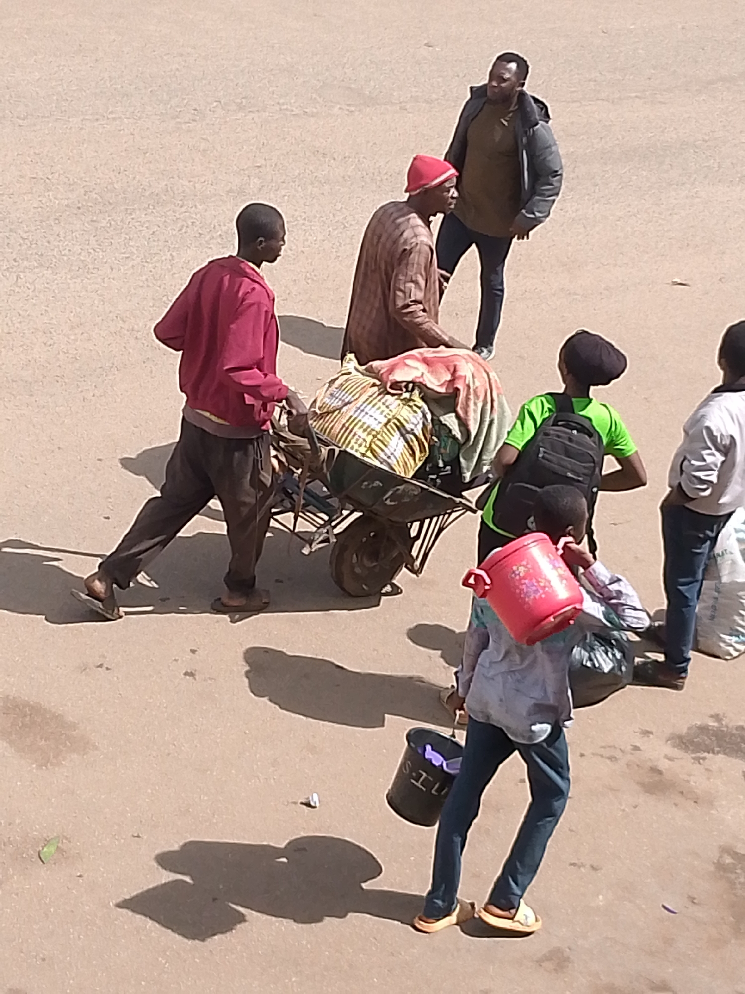 The image displays an outdoor scene in Bukuru, Nigeria, under direct sunlight, indicated by pronounced shadows cast on a light-colored, even ground surface.
In the mid-left, a dark-skinned male subject, wearing a red long-sleeved jacket, dark trousers, and dark flip-flops, is positioned with his right leg forward, seemingly engaging with a metal wheelbarrow. To his immediate right and behind the wheelbarrow, a second dark-skinned male subject, wearing a red cap and a light-brown patterned long-sleeved garment, stands. He is also interacting with the heavily laden wheelbarrow, which contains multiple bundled textiles, prominently featuring a yellow-striped fabric.
Standing to the right and slightly behind the wheelbarrow group, a dark-skinned male subject wears a dark grey puffer jacket over an olive green shirt, dark trousers, and sneakers. His posture is upright, and he observes the activity.
Further to the right, a subject with their back to the viewer is dressed in a neon green t-shirt, dark trousers, and a dark head covering, carrying a black backpack. Directly in front of and slightly below this individual, another subject in a light-colored patterned long-sleeved shirt and dark trousers carries a red plastic pot adorned with floral patterns in their left hand and a dark plastic bag in their right. This individual wears light-colored sandals.
On the far right, partially visible, a subject wearing a white jacket and dark trousers is seen holding a large white sack.
The ground surface is a consistent light tan color, and shadows are uniformly cast towards the lower-left portion of the frame. Small pieces of debris, including a green leaf and a white crumpled object, are visible on the ground.