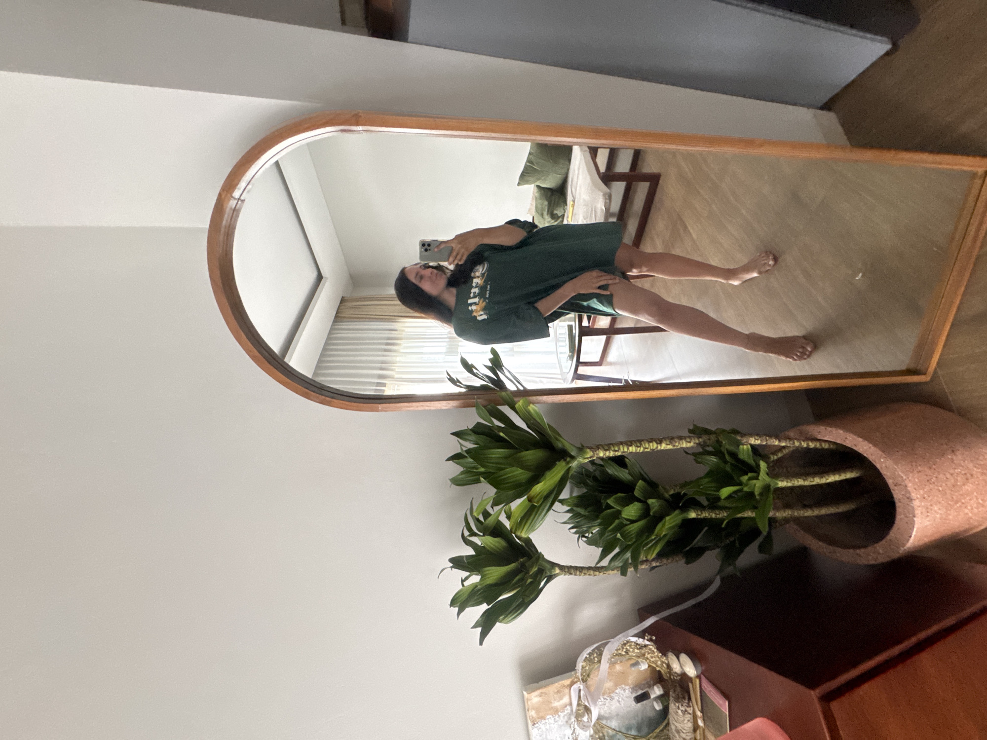 A young woman with dark hair is taking a mirror selfie in what appears to be a well-lit, contemporary living room in North Kuta, Indonesia. She is wearing an oversized, dark green t-shirt dress with a graphic print on the front and appears to be barefoot. The room features a large, arched wooden-framed mirror, through which the woman is visible. To the left of the mirror is a potted plant with lush green foliage. In the background, reflected in the mirror, is a sofa with green cushions, a coffee table, and curtains. The flooring is a light-colored wood.