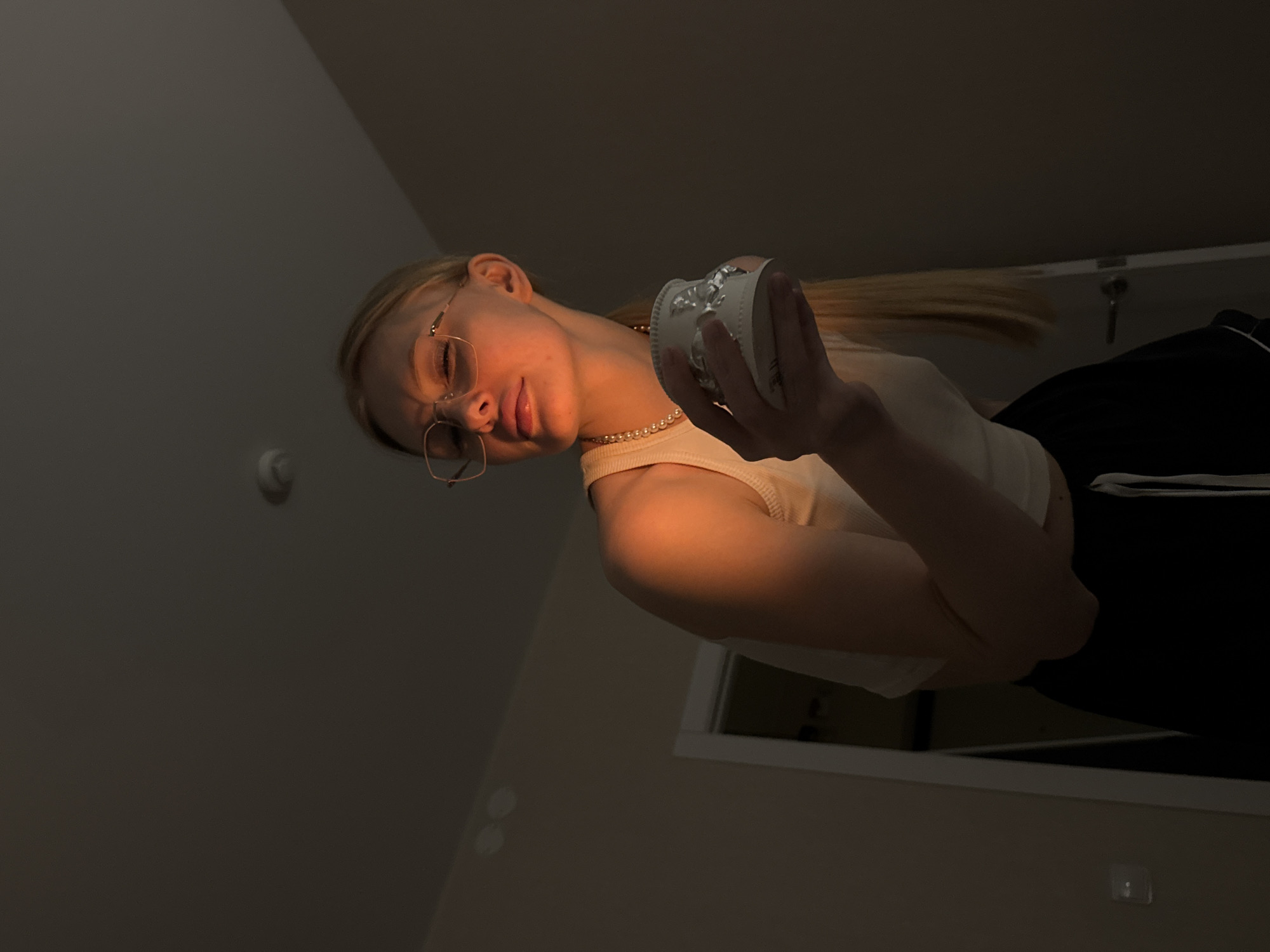 The image captures a young woman from a low-angle perspective, creating an intimate and unique viewpoint. She has blonde hair and is wearing round, light-rimmed glasses. A pearl necklace adorns her neck, complementing her light-colored top, possibly a tank top or camisole. She is also wearing dark bottoms, likely pants or shorts with a white stripe. In her right hand, she holds a small, decorative, light-colored object that appears to be an ornate cup, candle holder, or vase, with intricate detailing and possibly a reflective or metallic top.
The dominant feature of the image is the dramatic, warm lighting that illuminates the woman's face, neck, and right arm from below, casting a soft, golden glow. Her expression is calm and composed, with a slight, relaxed smile. The background is dark and indistinct, suggesting a dimly lit indoor setting, likely evening or night. Subtle architectural elements like a ceiling corner and a white square shape (possibly a window or door frame) are visible in the background, along with a white circular object on the wall/ceiling, which might be a smoke detector or light fixture. The overall mood is quiet and contemplative, emphasized by the soft, focused lighting. No text is visible in the image.