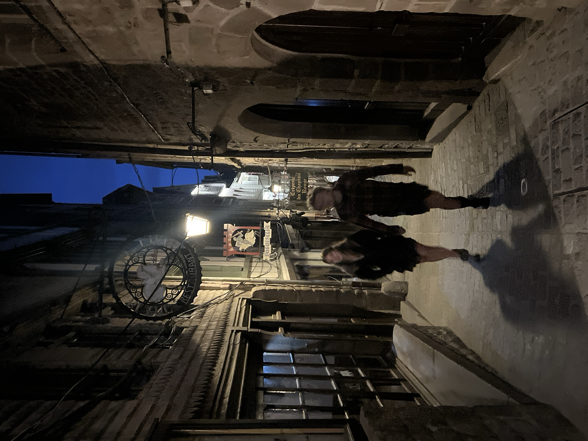 The image depicts a narrow, ancient cobblestone alleyway in Le Mont-Saint-Michel, France, at twilight or dawn. Two individuals, a female and a male, walk hand-in-hand towards the viewer on the uneven stone pavement. The female, positioned on the left, has visible light brown hair and wears dark attire, casting a prominent shadow behind her to the left. The male, on the right, wears a plaid shirt and dark shorts, also casting a discernible shadow to his right and behind him.
The alley is enclosed by tall, aged stone buildings on both sides. The building on the left features horizontal wooden siding on its upper facade and a distinctive sawtooth-patterned wooden overhang above a large, multi-paned window at ground level. Above this window, a large, ornate circular metal sign with decorative scrollwork is affixed to the building, displaying the word "RESTAURANT" at its bottom, but lacking clock hands. Further down the alley on the left, a rectangular red sign with a golden crest and the name "Duguesclin" is visible.
The building on the right is constructed of rough-hewn stone blocks, featuring two dark, arched doorways in the foreground and various wires and conduits running along its surface. Multiple traditional street lamps illuminate the scene with warm, artificial light, casting long shadows and highlighting architectural details. In the distance, another white rectangular sign reading "Crêperie du Chapeau Rouge" is discernible. A sliver of deep blue sky is visible above the narrow gap between the buildings at the top of the image. Manhole covers are embedded in the cobblestone pathway.