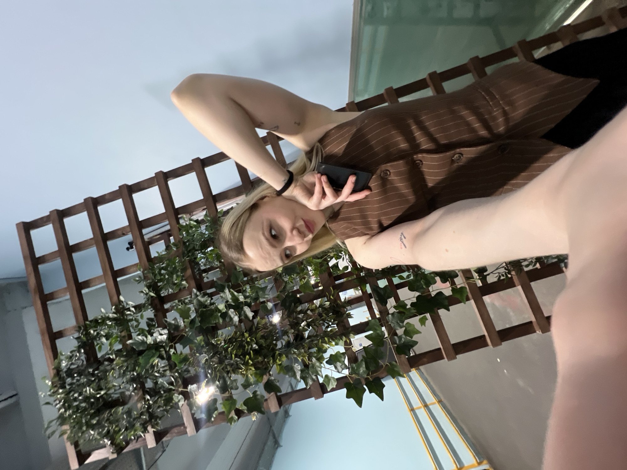 A waist-up, tilted, selfie-style photograph depicts a woman holding a black smartphone. Her face is oriented towards the camera. She is fair-skinned and blonde. She wears a brown vest with light brown vertical stripes. She has a black bracelet on one wrist. There are at least two tattoos visible on her arms. Behind her is a wooden trellis with climbing ivy. Behind the trellis is a pale blue wall and a partially visible structure with blue and yellow vertical stripes.
While not directly specified in the image, the metadata claims that this image was taken in Khimki, Russia.