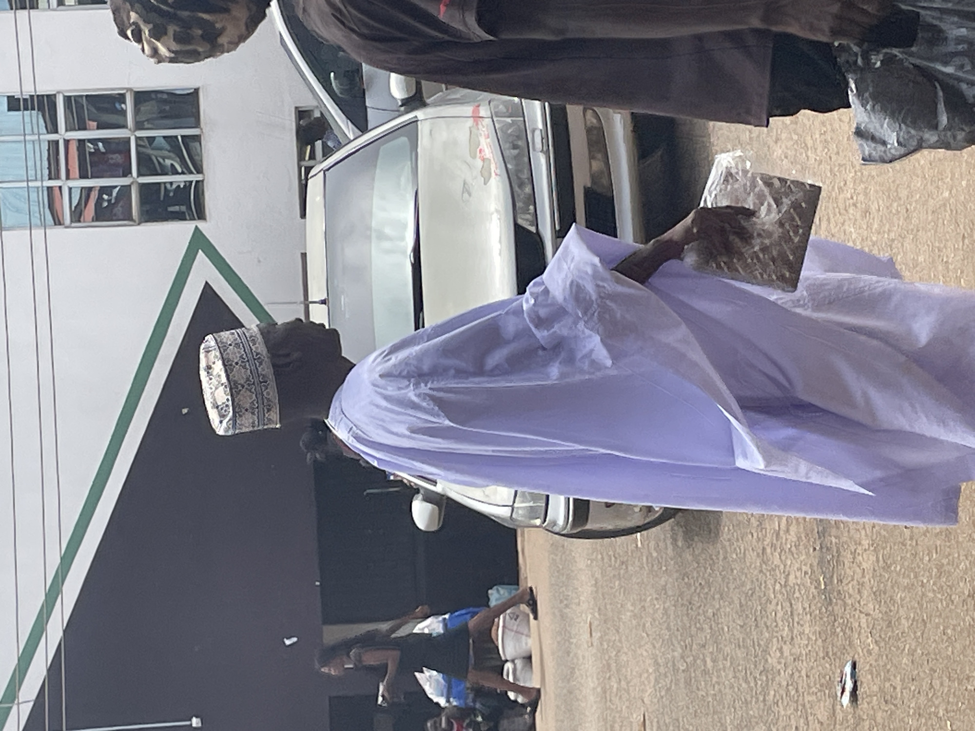 In the city of Bukuru, Nigeria, a man wearing a white traditional outfit and a patterned cap is captured mid-stride, holding a clear plastic bag filled with what appears to be food items. He is walking beside a silver car, his focus directed towards the left of the frame. In the background, a woman in a dark dress is seen walking away from the viewer, accompanied by piles of goods. The environment suggests a bustling urban setting with buildings and other vehicles present.