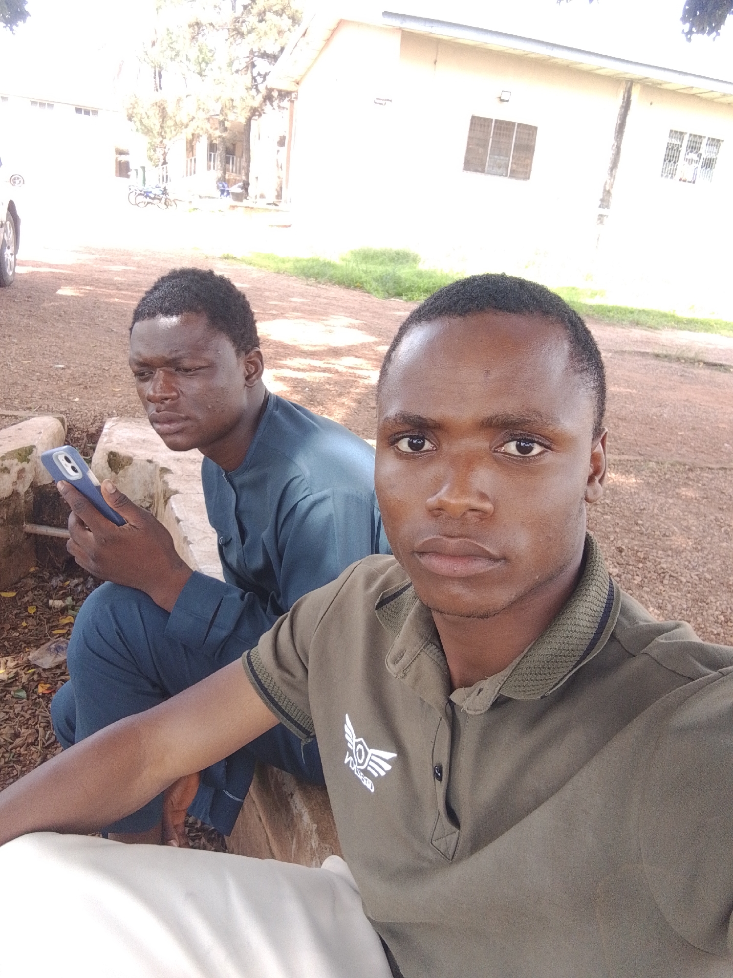 Two young Black men are outdoors, seated on a concrete ledge in front of a light-colored building with several windows. The man on the right is closer to the camera and is looking directly at it. He has short, dark hair, dark eyes, and is wearing a green polo shirt with a logo on the left chest. The man on the left is looking down at a blue smartphone he is holding in his left hand. He has short, dark hair and is wearing a dark blue, collared shirt. The ground around them is covered in dirt and loose gravel, with some dry leaves scattered about. In the background, there are trees and a white building with a corrugated metal roof. A motorcycle is visible parked in front of the building. The overall lighting suggests it is daytime.