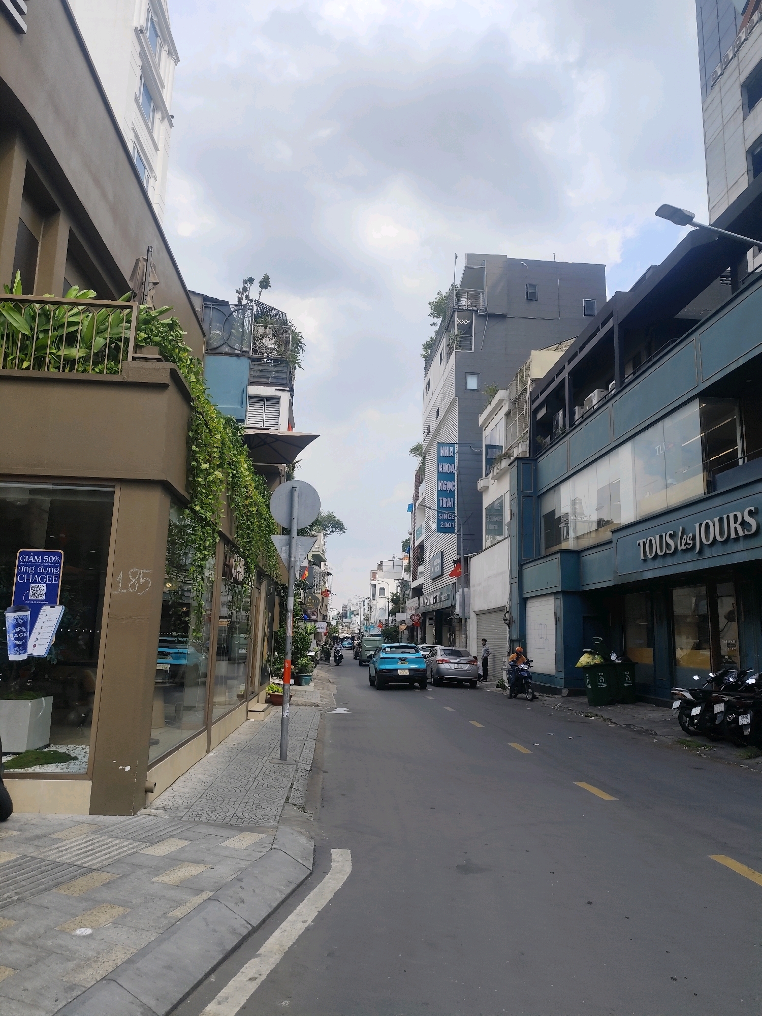 The image shows a street scene in Ho Chi Minh City, Vietnam, likely in an urban commercial area. The perspective is from a sidewalk looking down a street lined with multi-story buildings. On the left, a brown building with large glass windows is prominent, featuring green plants cascading down its facade and a sign advertising "CHAGEE" with a discount. Further down the street, there are more buildings, some with blue and grey facades, and signage for businesses like "TOUS les JOURS".
The street itself is paved with asphalt and marked with yellow lines. Several vehicles are present, including blue and silver cars, and numerous motorcycles, some of which are parked along the sidewalk. A person is seen riding a motorcycle and another individual stands near a silver car. Two green trash bins are situated on the sidewalk. The sky is overcast with clouds. The architecture suggests a mix of modern and older styles, common in Vietnamese cities. The presence of Vietnamese signage confirms the location.