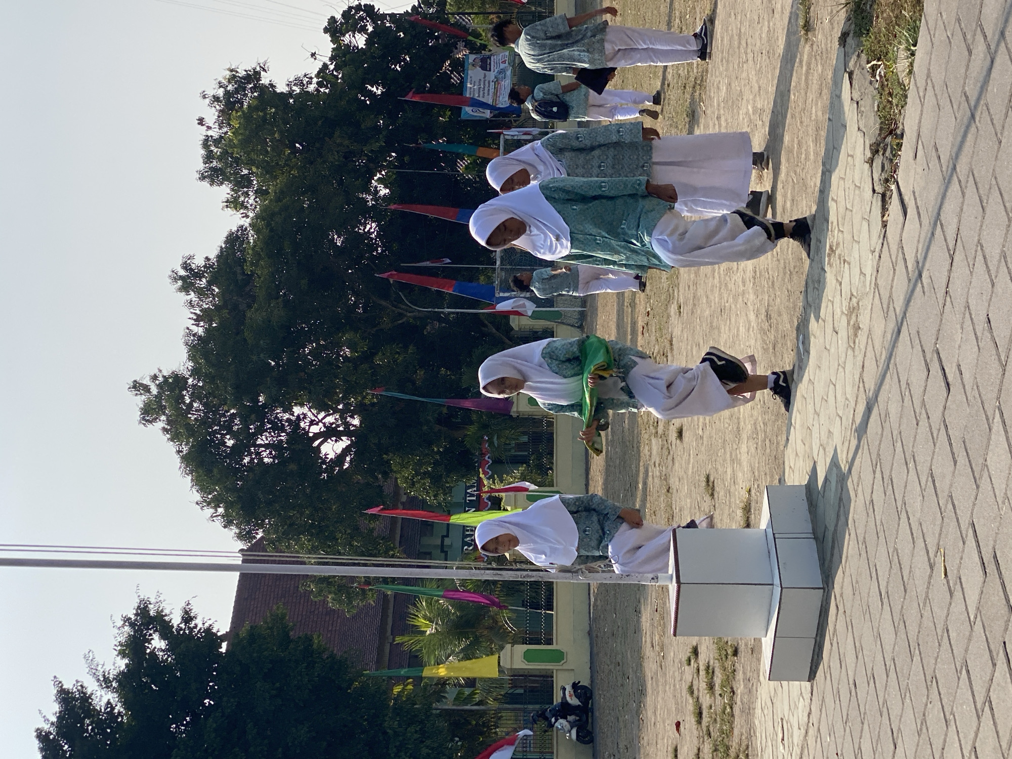 In Kasihan, Indonesia, a group of students wearing hijabs and matching teal and white uniforms are participating in a flag-raising ceremony. Four girls are prominently featured in the foreground, marching in formation. One girl on the left is holding a green object, possibly a flag or banner, as she walks. Another girl next to her is also holding a similar green item. The other two girls in the center are walking forward, looking ahead. In the background, several other students in similar uniforms are visible, some with backpacks. Colorful flags, including red and white Indonesian flags, are displayed on poles behind the students, along with a building with the text "RAHAN TAM" visible on its facade. The scene takes place outdoors on a dirt field adjacent to a paved walkway. The sky is clear and bright, indicating a sunny day.