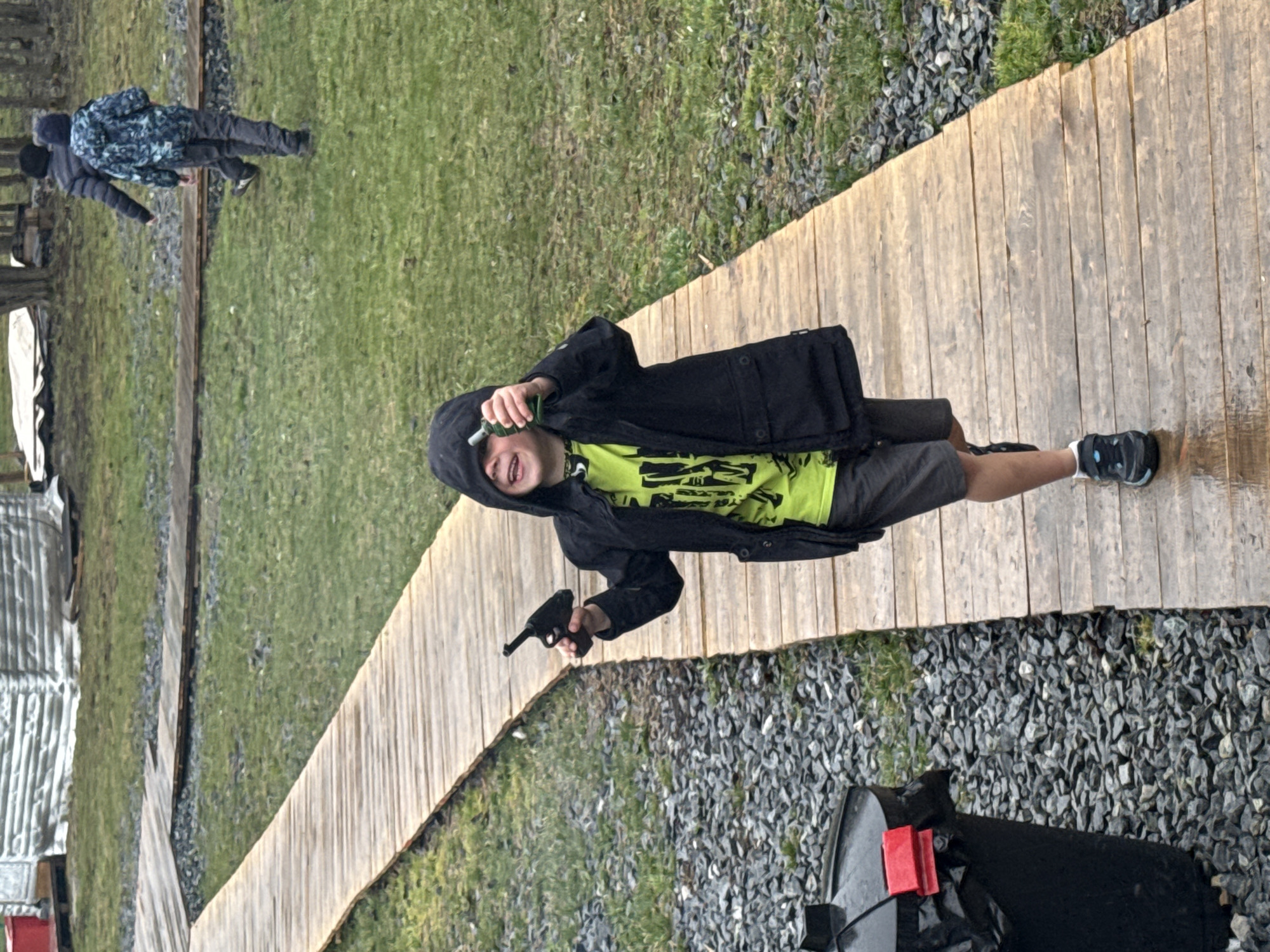 The image captures an outdoor scene in Yekaterinburg, Russia, featuring a young boy in the foreground and two other individuals in the background. The central subject is a boy, appearing to be of elementary school age, wearing a black hooded jacket over a lime-green t-shirt with a dark graphic design, gray shorts, and dark athletic shoes. He holds a black toy pistol in his left hand, pointing it downwards, and a small green plastic bottle in his raised right hand. His mouth is open, suggesting he is laughing, shouting, or expressing excitement.
The boy stands on a wooden plank pathway that stretches across the scene. To his right, the ground is covered in gray gravel, and further beyond are green grassy areas. In the mid-background, two other people are visible, standing near what appear to be train tracks. One wears a patterned or camouflage-style jacket, and the other a dark hooded jacket. On the far left, a corrugated metal structure is partially visible. The lighting indicates it is daytime, with soft, diffused light suggesting an overcast sky. The setting appears to be a recreational or semi-wild outdoor area, possibly near a disused railway line. No visible text is clearly discernible on any of the subjects or objects.