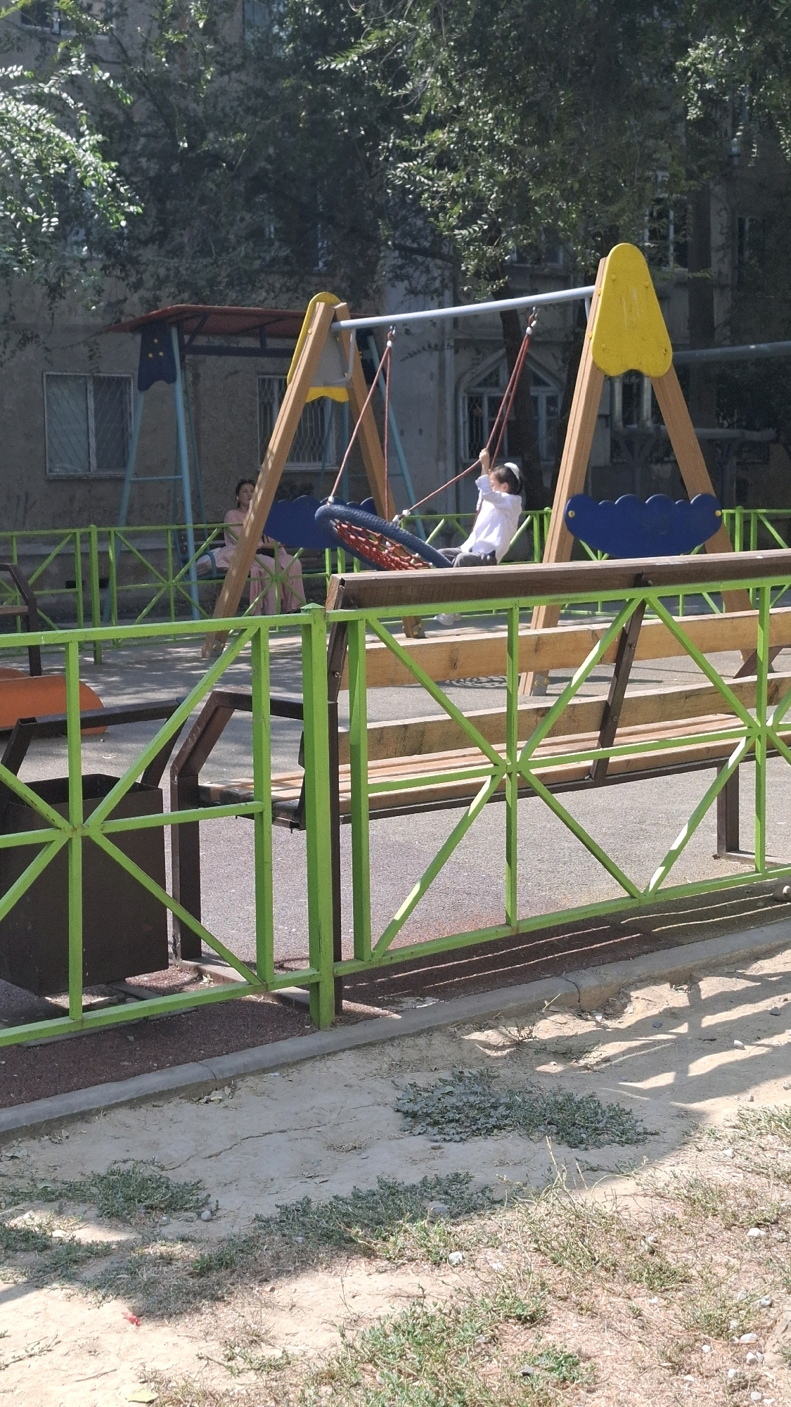 An outdoor public playground in Shymkent, Kazakhstan, features a swing set with a blue, web-like seat and a child in a white shirt and dark pants swinging. To the left of the swing set, a woman in a pink dress is seated on a bench. The playground is enclosed by a green metal fence with a wooden bench inside. The ground is covered with sand and sparse vegetation. In the background, a building with windows and trees is visible.