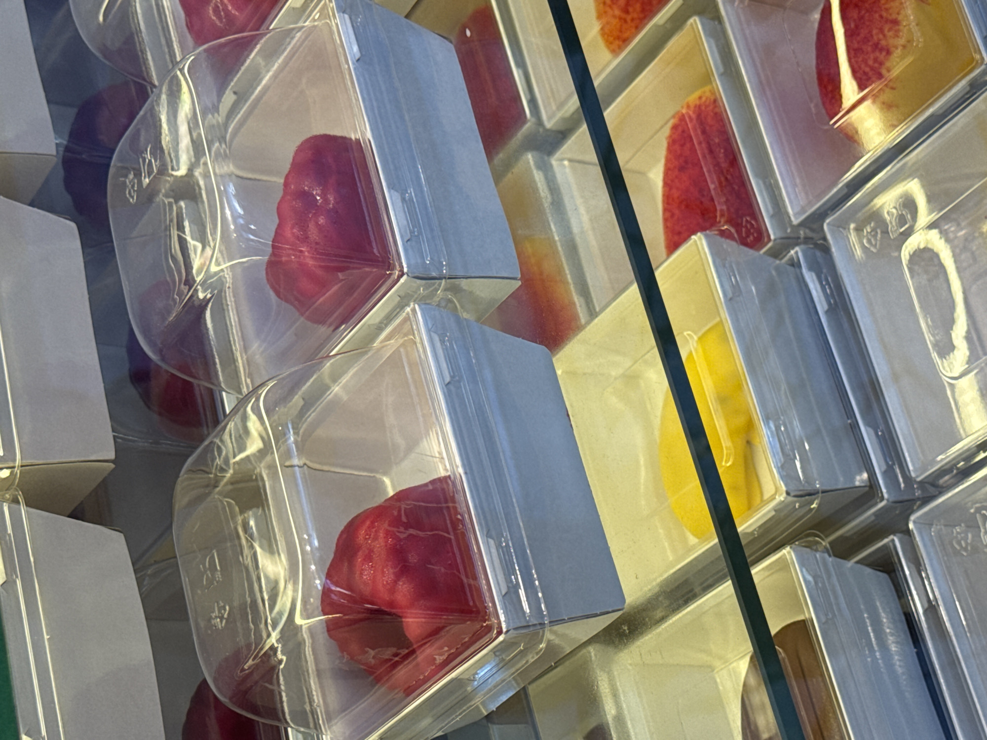 The image shows a close-up, eye-level view of a refrigerated display case filled with individually packaged desserts. The desserts appear to be small cakes or pastries with a glossy, red, heart-shaped topping. They are housed in clear plastic dome containers, which are placed inside light grey square cardboard sleeves. Some of the desserts have a slightly bumpy texture on their surface. Below the top layer of red desserts, there are visible trays containing yellow and red desserts, also in similar packaging. The overall environment suggests a food establishment, possibly a bakery or dessert shop. There are no people visible in the image. The lighting is bright and likely artificial, reflecting off the plastic packaging and the shiny dessert toppings. The context of Sochi, Russia, is not visually evident in the provided image.